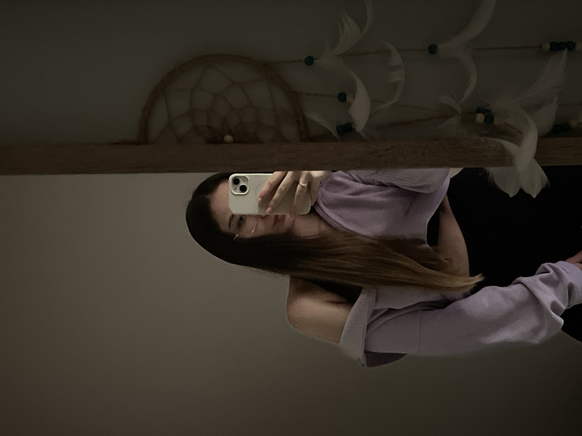 A woman is taking a selfie in a mirror. She is wearing a light purple sweatshirt and holding a white smartphone. The mirror is on a shelf, and above the shelf, there is a dreamcatcher and other decorative items. The location is Unknown, Unknown.