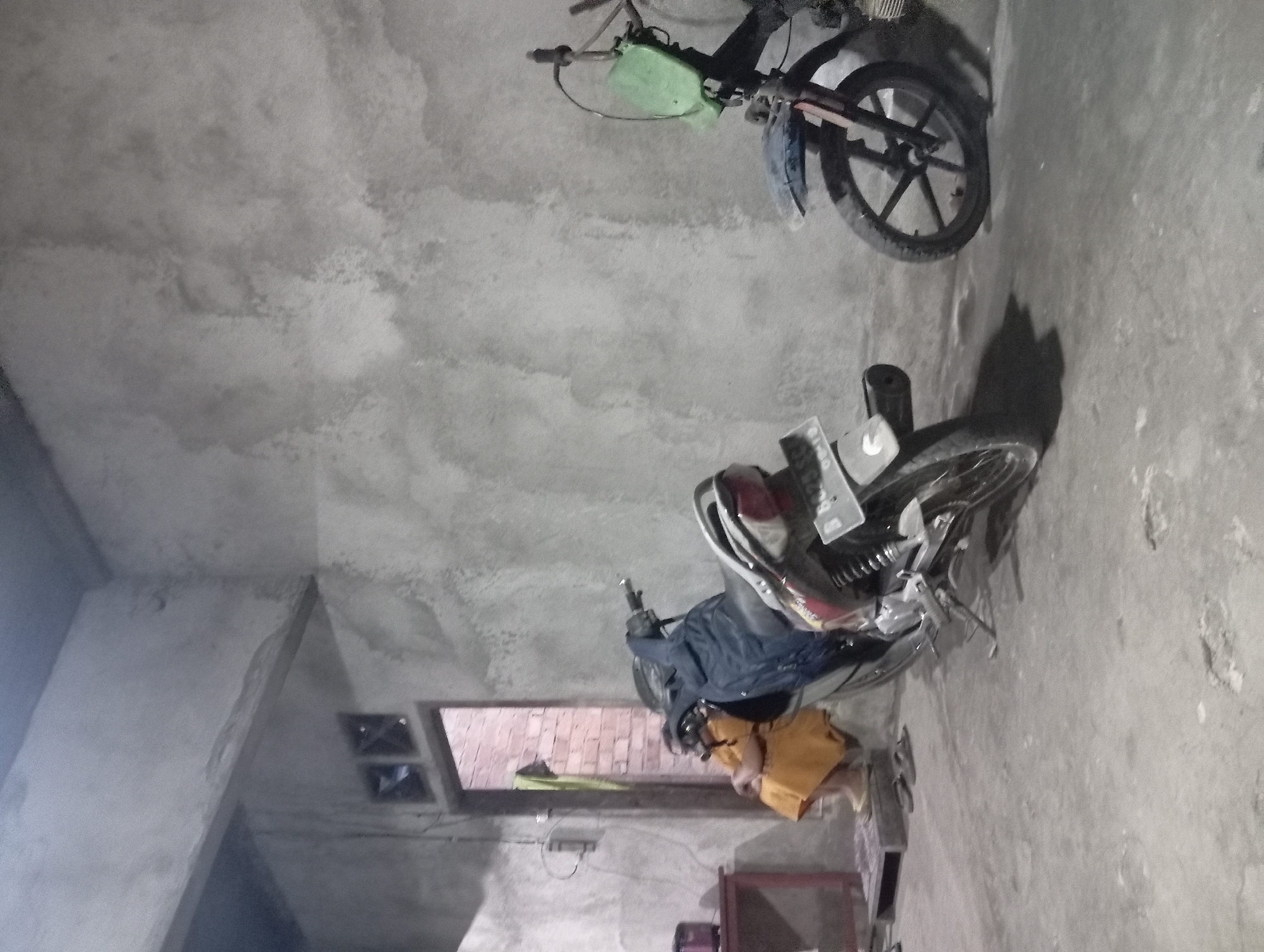 The image depicts an indoor scene, possibly a garage or an open-air shelter, in Rejosari, Indonesia. The environment is characterized by rough concrete walls and a concrete floor. In the foreground, two motorcycles are parked. The motorcycle on the left is partially visible, showing its rear wheel, fender, and license plate which reads "B 6028...". It appears to be a dark-colored motorcycle with some red accents. The motorcycle on the right is more prominently displayed, featuring a black frame, a blue fender, and a green plastic shield attached to the handlebars. Its front wheel, suspension, and part of the engine are visible.
To the left of the motorcycles, a person is seated, facing away from the viewer and towards an open doorway. The person is wearing an orange-colored garment and appears to be relaxed or resting. The doorway leads to an area with exposed brick walls. Near the seated person, there is a red table with some objects on it, including what looks like a small appliance, possibly a rice cooker. A pair of sandals is placed on the floor near the table. The lighting in the scene is dim, suggesting it might be evening or an indoor space with limited light sources. The overall impression is of a functional, utilitarian space.