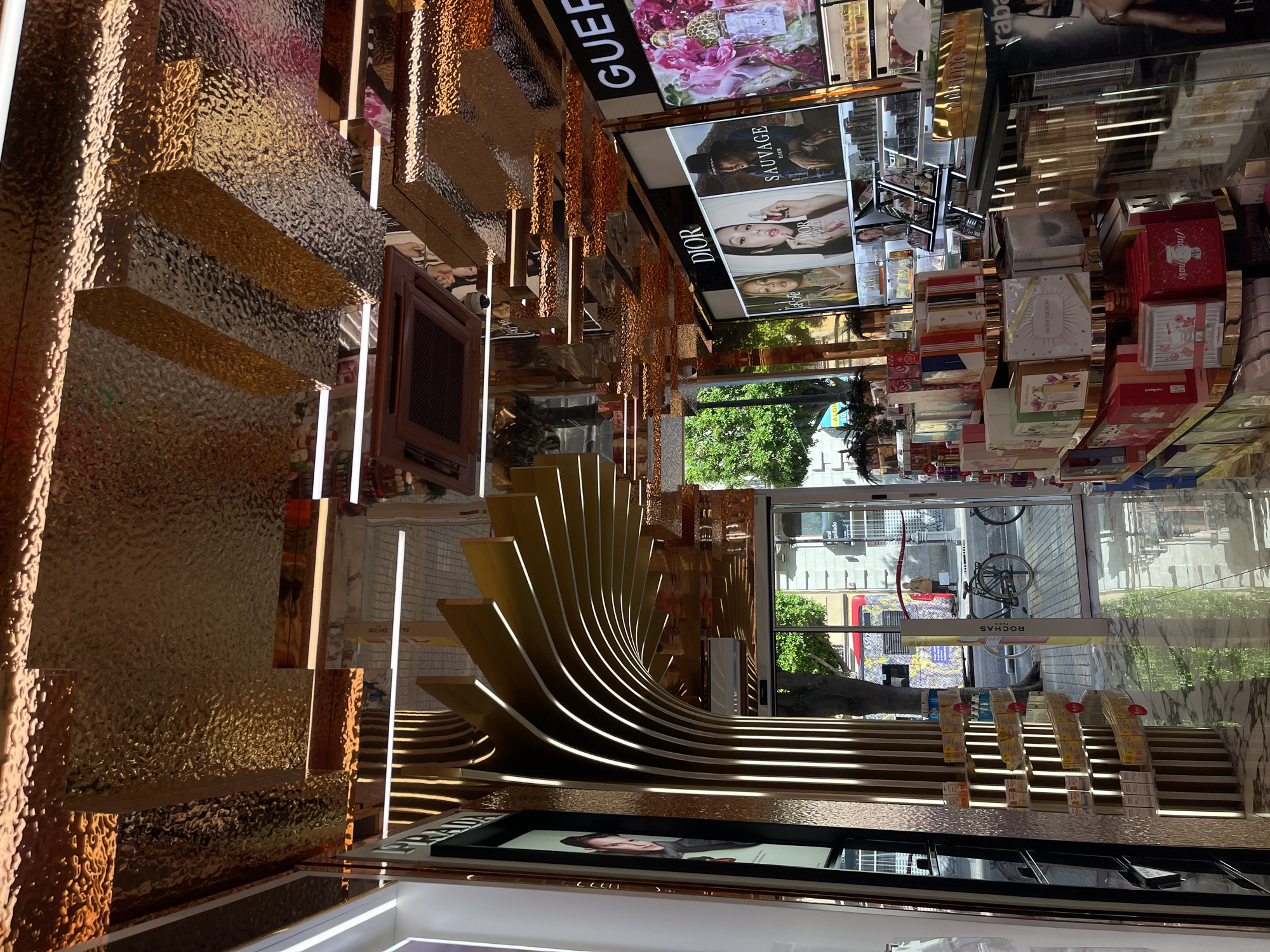 This image provides a detailed view of the interior of a high-end perfume and cosmetics store located in Valencia, Spain. The scene is brightly illuminated by various light sources, creating a luxurious and reflective environment.
The ceiling is a prominent architectural feature, composed of multiple layered panels of textured, hammered metal with a reflective bronze-gold finish. These panels create a fragmented, three-dimensional effect, reflecting linear LED strip lights, digital displays, and other store elements. A rectangular air conditioning vent is centrally recessed into one of the ceiling panels.
On the left side of the store, a large vertical digital display screen showcases a woman's profile, with the brand name "PRADA" visible at the top. Adjacent to this display is a unique curved wall installation featuring numerous vertical, gold-toned slats that arc inward, each embedded with linear LED lighting, creating a luminous, ribbed pattern.
On the right side, multiple large digital display screens are mounted, advertising luxury perfume brands. One set of screens prominently displays "DIOR," featuring three separate images: two female models and one male model advertising "SAUVAGE Elixir." Further right, another screen advertises "GUERLAIN" with a floral motif. Below these screens, numerous packaged perfume and cosmetic products are displayed on tiered, curved shelves and glass-fronted counters, with brands such as "BOUCHERON" and "ROCHAS" identifiable. A vertical "ROCHAS PARIS" sign post stands near the store entrance. A partial advertisement for "Rabanne" (Paco Rabanne) is visible on the far right.
The floor is made of polished white marble or tile, reflecting the ceiling, lighting, and product displays. Through a large glass entrance door and adjacent window, an urban street scene is visible. Outside, there are green trees, stone building facades, a red and yellow public bus displaying the number "6502" and graphic art, a distant person in a light-colored coat, and a black bicycle parked on the sidewalk. Reflections of the store's interior are also visible on the glass surfaces. Faint reflections of people are subtly present on the reflective ceiling panels.
