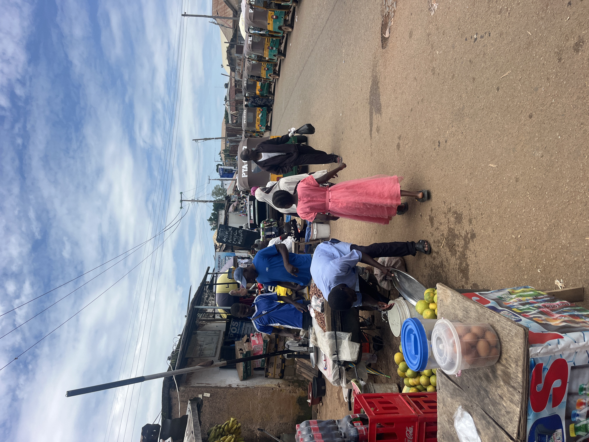 The image depicts a bustling street market scene in Gura Top, Nigeria. In the foreground, a stall displays a variety of goods, including a container of eggs and a stack of red crates filled with bottled drinks. Several individuals are engaged in transactions and moving about the market. On the left, two men in blue shirts are interacting at a stall laden with produce, possibly nuts and fruits. A third man, wearing a light blue shirt and dark pants, is bent over, attending to something on the ground. Further down the street, a young girl in a pink dress walks away from the camera, followed by an adult. A line of auto-rickshaws, some bearing the "PTA" logo, are parked along the right side of the road, indicating a transportation hub within the market. The environment suggests a warm, possibly humid, day with a cloudy sky overhead. The structures visible are simple, with corrugated metal roofs and unpainted walls, characteristic of informal markets in many Nigerian cities. The overall impression is one of lively commerce and daily life in a vibrant urban setting.