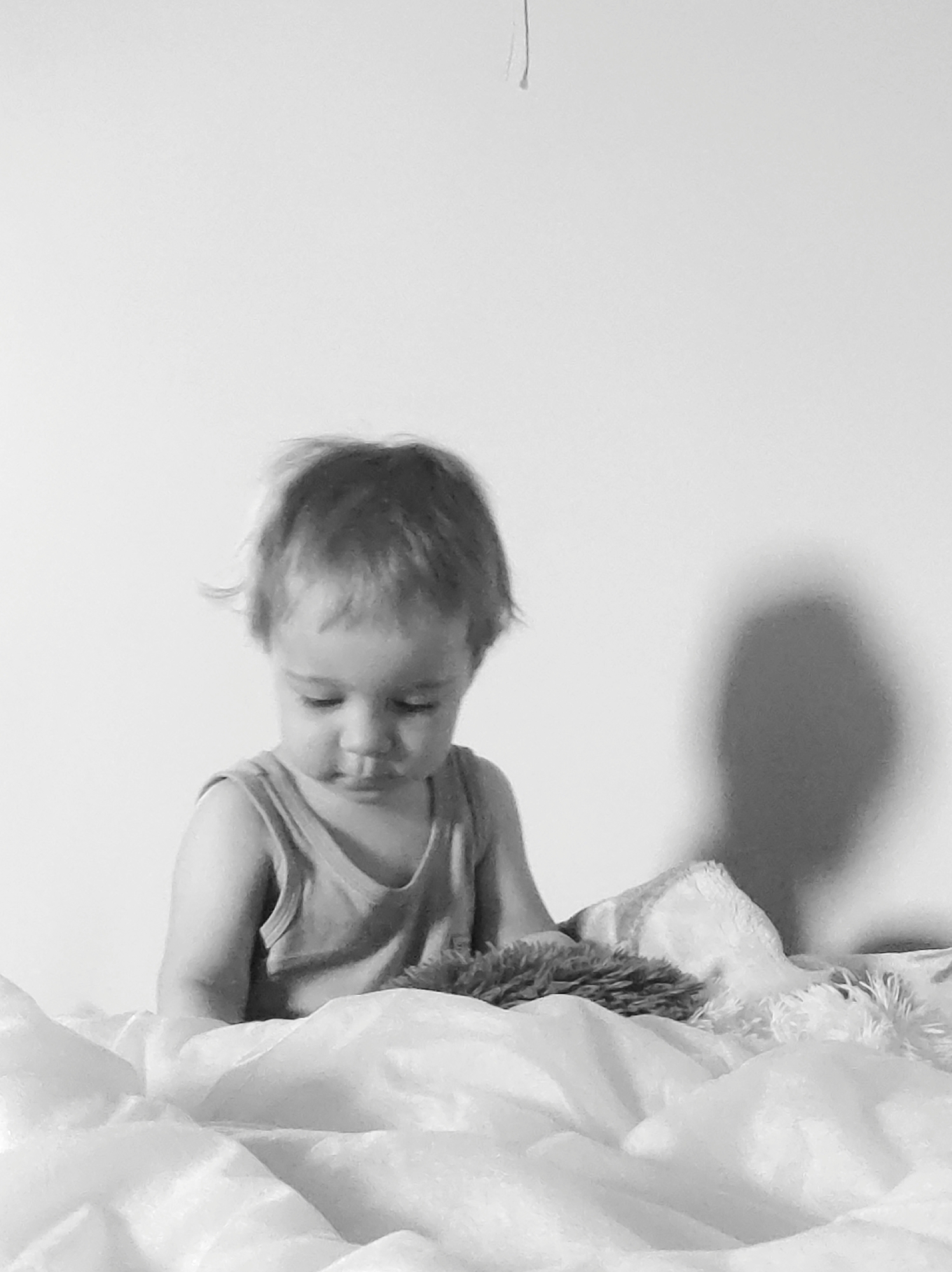 A black and white close-up depicts a young child sitting in a bed covered with a white, rumpled blanket or sheet. The child is wearing a sleeveless top and appears to be looking down, possibly at a fluffy toy or object on the bed. A shadow is cast on the plain white wall behind the child. The location is unknown.