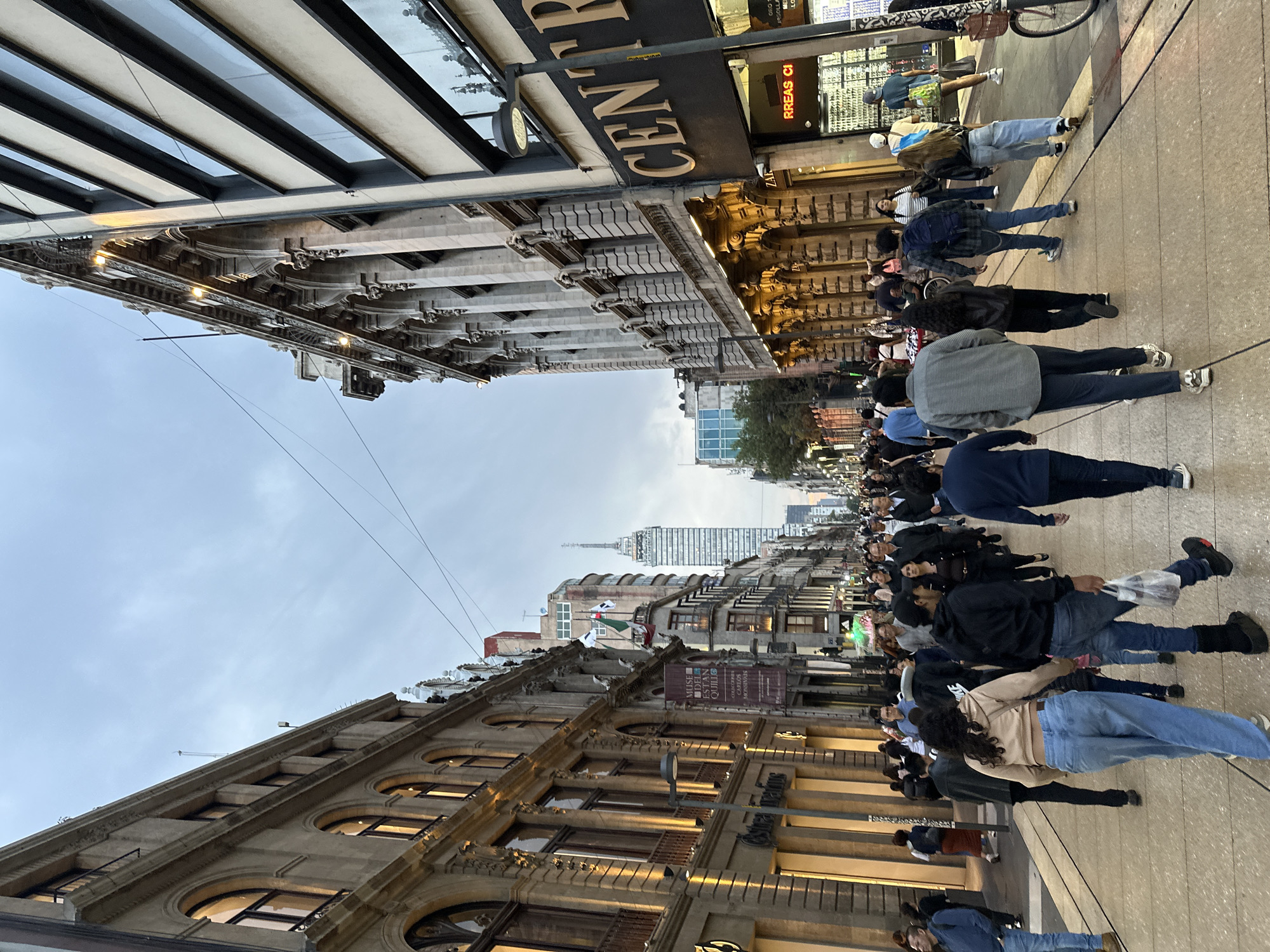 The image depicts a street scene in Mexico City, Mexico. The pedestrian walkway is densely populated with people walking in both directions. On the right side of the frame, there is a building with a sign that reads "CENTRO." On the left side, the architecture of the buildings features arched windows and ornate details. In the background, a tall skyscraper is visible, piercing the cloudy sky. Electrical wires are strung overhead. The overall environment suggests a bustling urban area.
