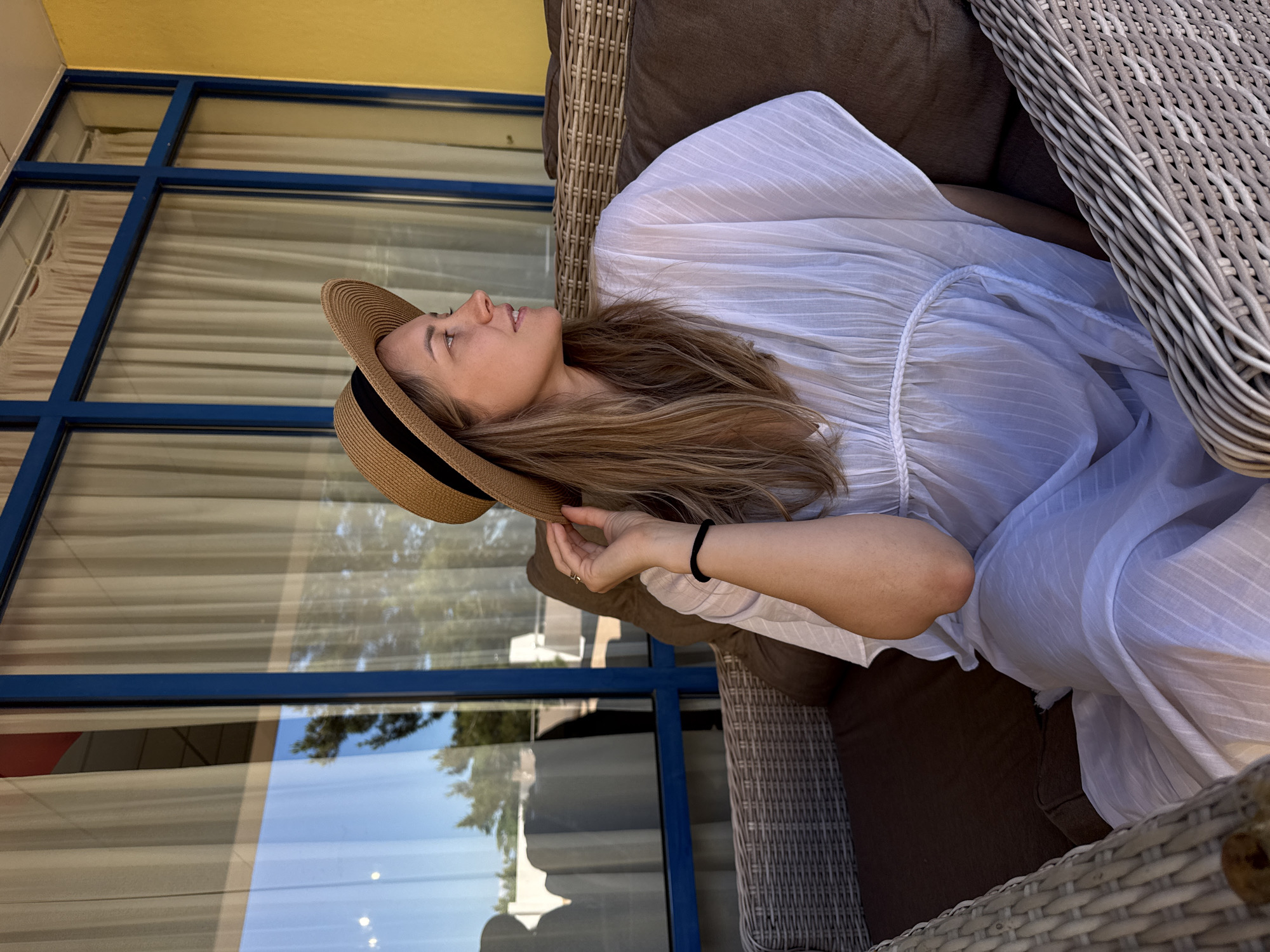 A young woman wearing a white dress and a straw hat is sitting on a wicker chair, looking to the right. She is sitting on a sofa, likely outdoors, with a blue window frame in the background. The window reflects trees and a building. The woman is holding her hat with her left hand. The image is taken during the day, with natural lighting. The location is not specified in the image, but the prompt requests it to be set in Nebug, Russia.