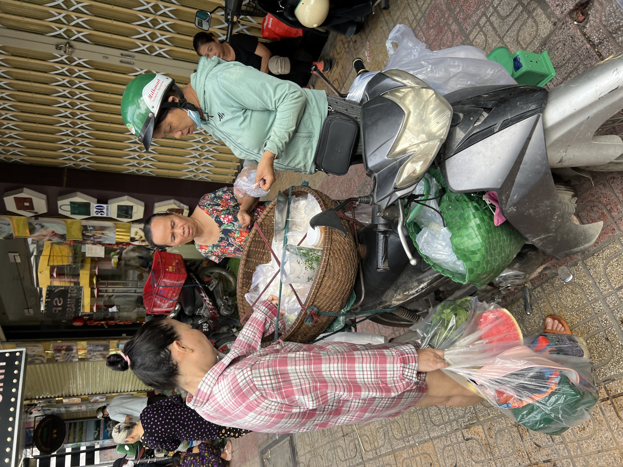 The image depicts a street scene in Nha Trang, Vietnam, likely a market or a bustling sidewalk. Three adult Vietnamese women are the central figures. On the left, a woman in a red and white plaid shirt is holding a large plastic bag filled with groceries, including watermelon slices. She appears to be interacting with a woman in the center who is wearing a floral print top and looking down. The woman on the right, wearing a green hooded jacket and a green helmet, is holding a plastic bag and appears to be exchanging items with the woman in the center. Both women are standing next to a motorcycle, which is parked on the sidewalk and laden with a woven basket containing more goods. The basket has white contents, possibly food items, wrapped in plastic. A green basket is also visible near the motorcycle's rear. In the background, there are storefronts with metal shutters and various signs and displays, typical of a commercial area. Another woman is visible in the background to the right, wearing a black top. The ground is paved with interlocking tiles, and a green scale is positioned near the motorcycle. The overall impression is one of everyday commerce and activity on a street in Vietnam.