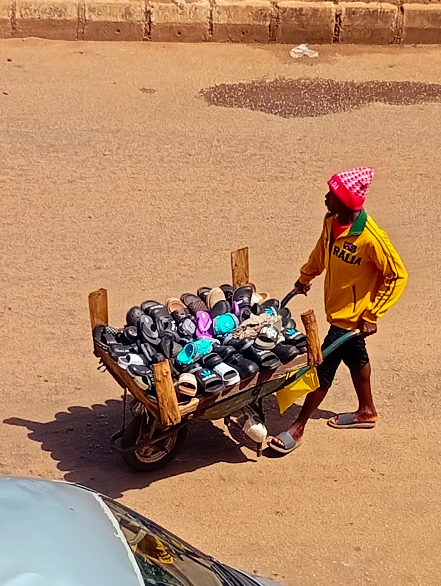 In Bukuru, Nigeria, a young Black male, wearing a pink and white checkered beanie, a yellow jacket with "RALIA" printed on the back, and black shorts, pushes a wheelbarrow laden with an assortment of colorful flip-flops and sandals. The wheelbarrow, constructed of wood and metal, has a single wheel and two wooden handles, one of which the man is gripping. The ground is a dusty, light brown surface, with scattered patches of darker dirt and a few small, dark objects. A blurred, light-colored car is partially visible in the foreground at the bottom left of the image. The background features a rough, textured wall made of rectangular blocks. A crumpled white plastic bag is visible near the wall in the upper center of the image. The scene appears to be an outdoor market or street.