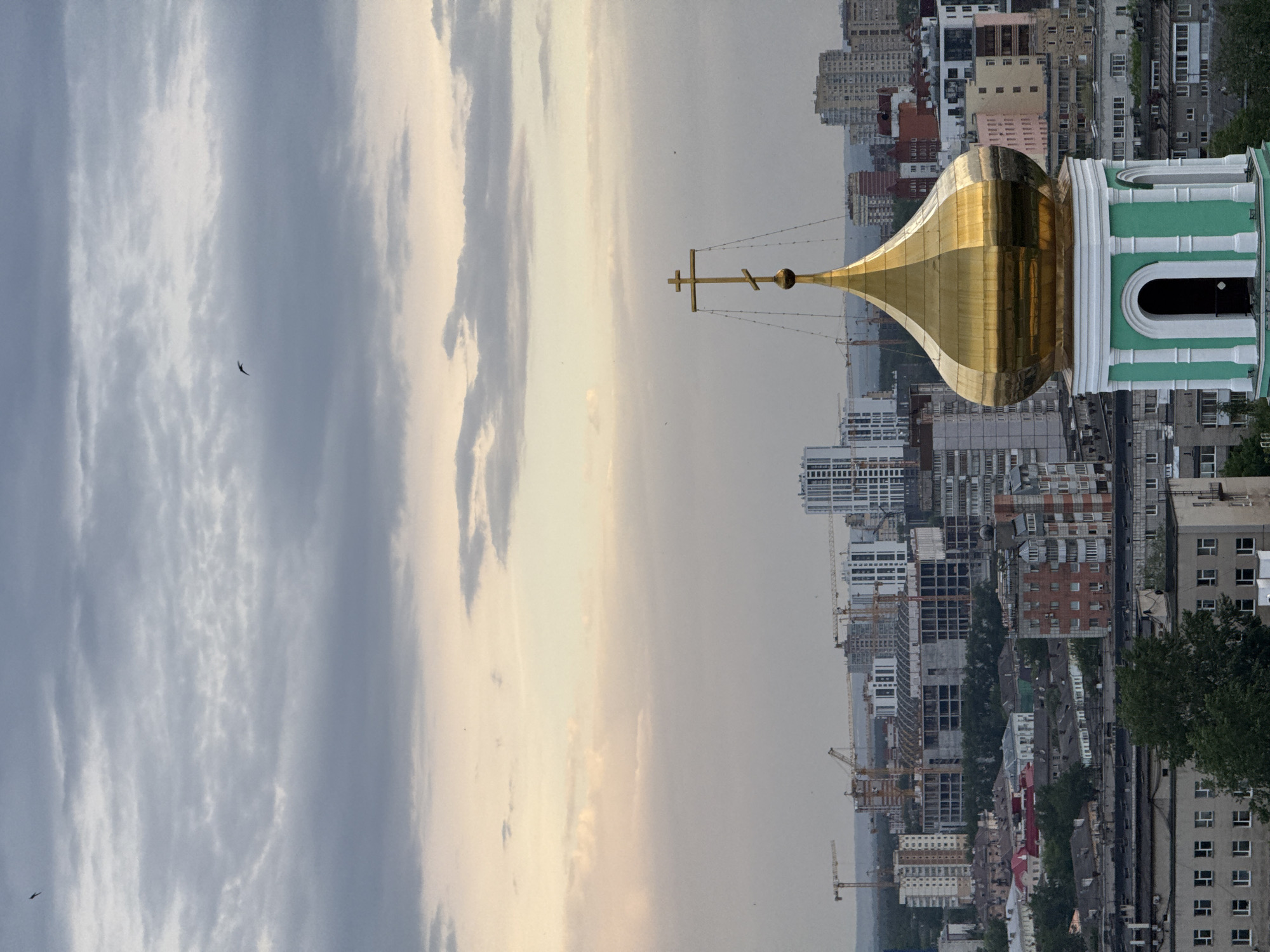 The image captures a view of Perm, Russia, with a prominent golden-domed church in the foreground. The church's dome is topped with a golden cross. In the background is a mix of buildings, including modern high-rises and older structures. Construction cranes are visible among the buildings, indicating ongoing development in the city. The sky is overcast with shades of grey and white clouds. A bird is flying in the sky. The image appears to be taken from an elevated perspective, possibly a rooftop or a high point in the city.