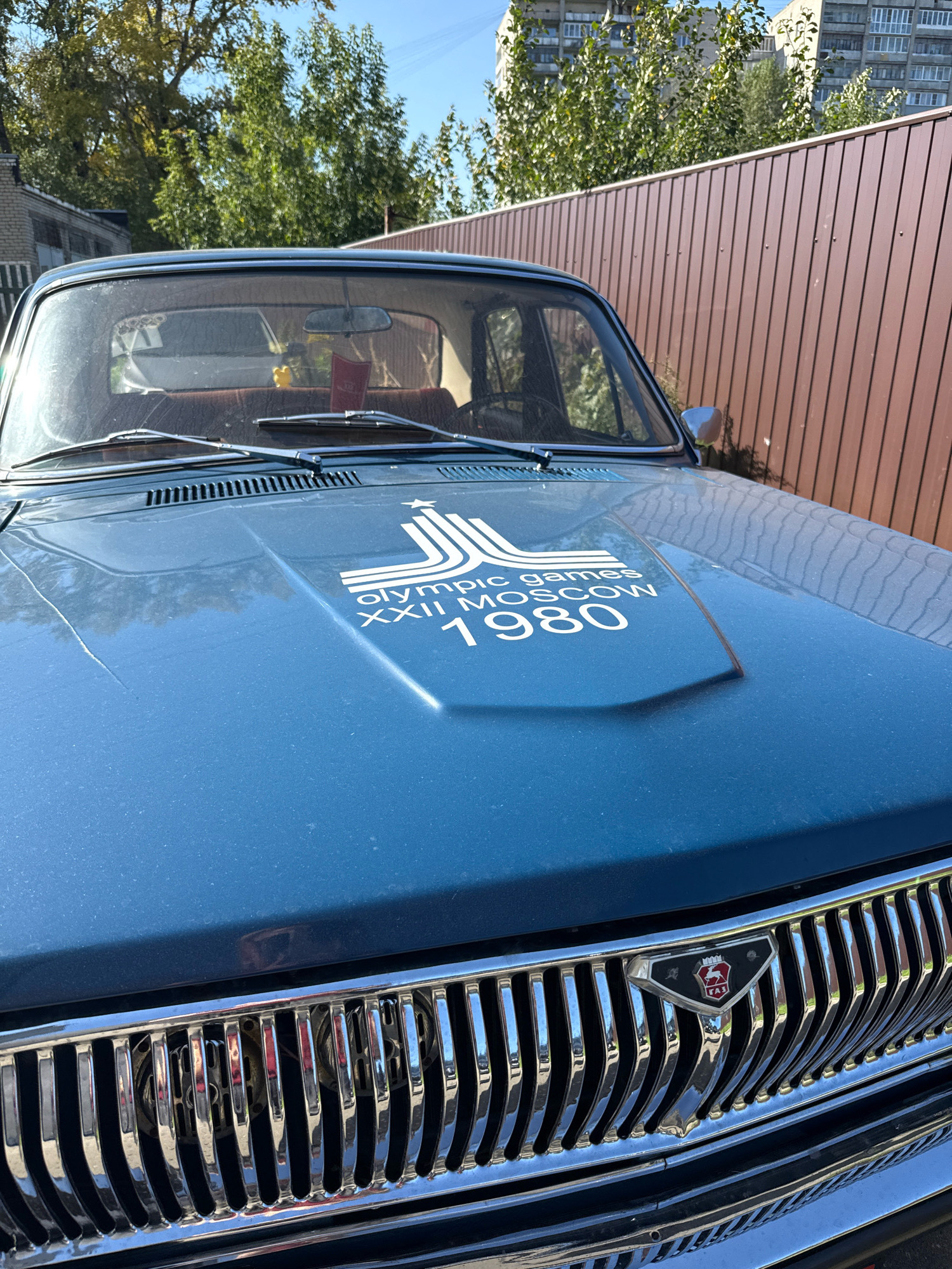 A close-up, eye-level exterior shot shows the hood and grille of a blue GAZ-24 "Volga" sedan parked outdoors on a sunny day. The car's hood is emblazoned with a white graphic depicting the Moscow 1980 Olympic Games logo and the text "OLYMPIC GAMES XXII MOSCOW 1980." The chrome grille of the car is visible at the bottom of the frame, featuring vertical bars and a triangular emblem with the GAZ logo. In the background, a tall brown wooden fence runs horizontally, with trees and a multi-story apartment building visible beyond it under a clear blue sky. The scene is set in Rusanovo, Russia. No people are present.