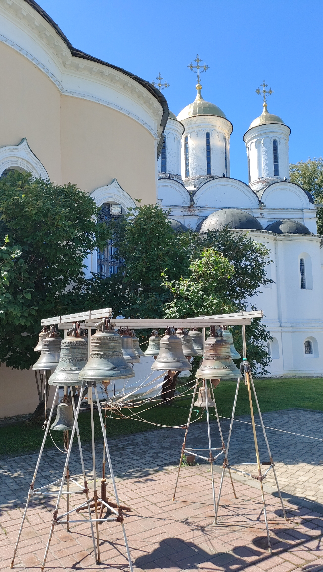 The image depicts a collection of large, bronze bells displayed outdoors on metal stands in front of a white and gold domed church. The church has multiple domes with golden crosses and arched windows. To the left of the church is a building with a curved, light yellow facade, also featuring arched windows. Green trees and foliage are visible between the buildings and around the area. The ground is paved with rectangular bricks. The scene is illuminated by bright sunlight, casting shadows. The bells are suspended from a horizontal metal frame supported by tripod-like structures. The overall setting suggests a religious or historical site. The church appears to be the Spaso-Preobrazhensky Monastery in Yaroslavl, Russia, known for its historic bell tower and collection of bells.