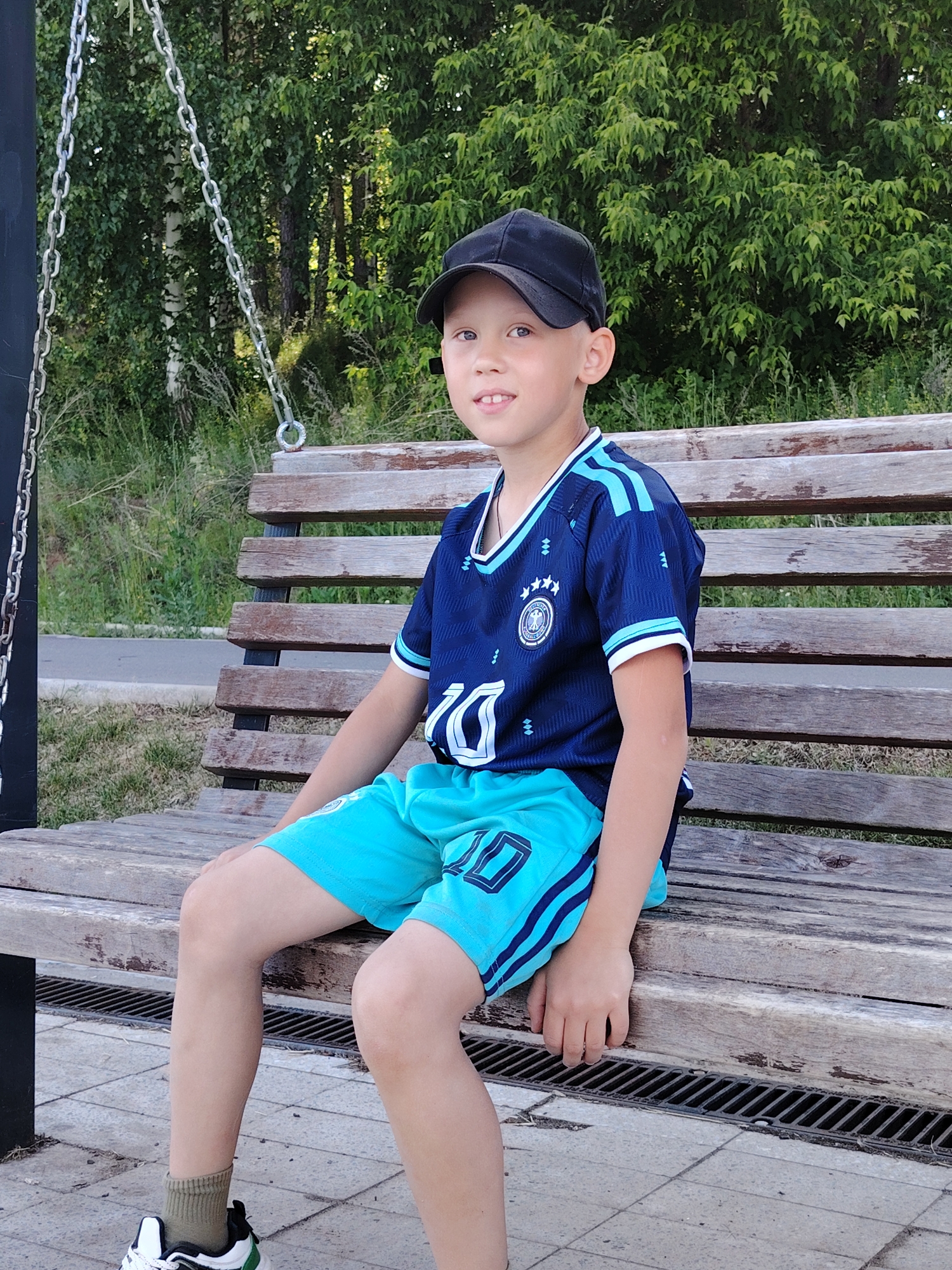 A young boy with short, light hair and blue eyes sits smiling on a wooden bench outdoors. He is wearing a dark blue sports jersey with light blue accents, light blue shorts with dark blue stripes, and a black baseball cap. His right hand rests on his leg, while his left hand is visible near his hip. He is also wearing olive green socks and white and green athletic shoes. The background features lush green foliage, suggesting a park or wooded area. To the left, a dark metal structure with chains is visible, possibly part of a swing set or playground equipment. The wooden bench he is sitting on appears worn. The ground in the foreground is paved with light-colored tiles and includes a dark metal grate. The lighting suggests it is daytime. The boy appears happy and relaxed.