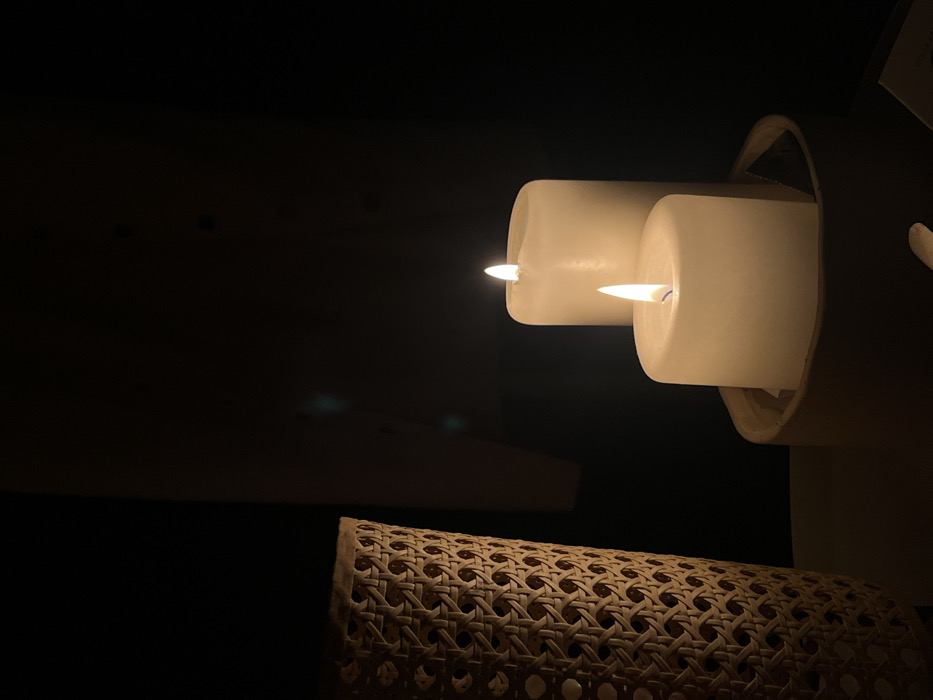 Two white candles, alight with small flames, sit atop a light wicker surface in a dimly lit room. The candles are partially enclosed in a white, circular holder. A dark shelf or table edge is visible in the background, contributing to the overall low-light atmosphere. The woven pattern of the wicker adds texture, contrasting with the smooth, solid forms of the candles. The scene evokes a sense of warmth and tranquility.