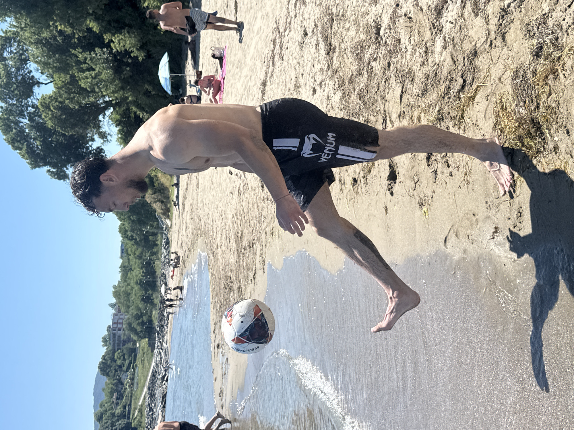 The image shows a man playing soccer on a sandy beach, with the sea visible to his left. The man is shirtless and wearing black shorts with white stripes and a logo that says "VENUM". He is in mid-air, kicking a soccer ball. The beach is populated with other people, some of whom are visible in the background, relaxing and enjoying the sunny weather. Lush green trees are visible behind the beach, and a building complex can be seen in the distance. The water is clear and shallow near the shore, with small waves breaking on the sand. The scene suggests a warm, sunny day, likely in the afternoon, given the length of the shadows. The overall mood is relaxed and recreational. The visible text on the man's shorts reads "VENUM".