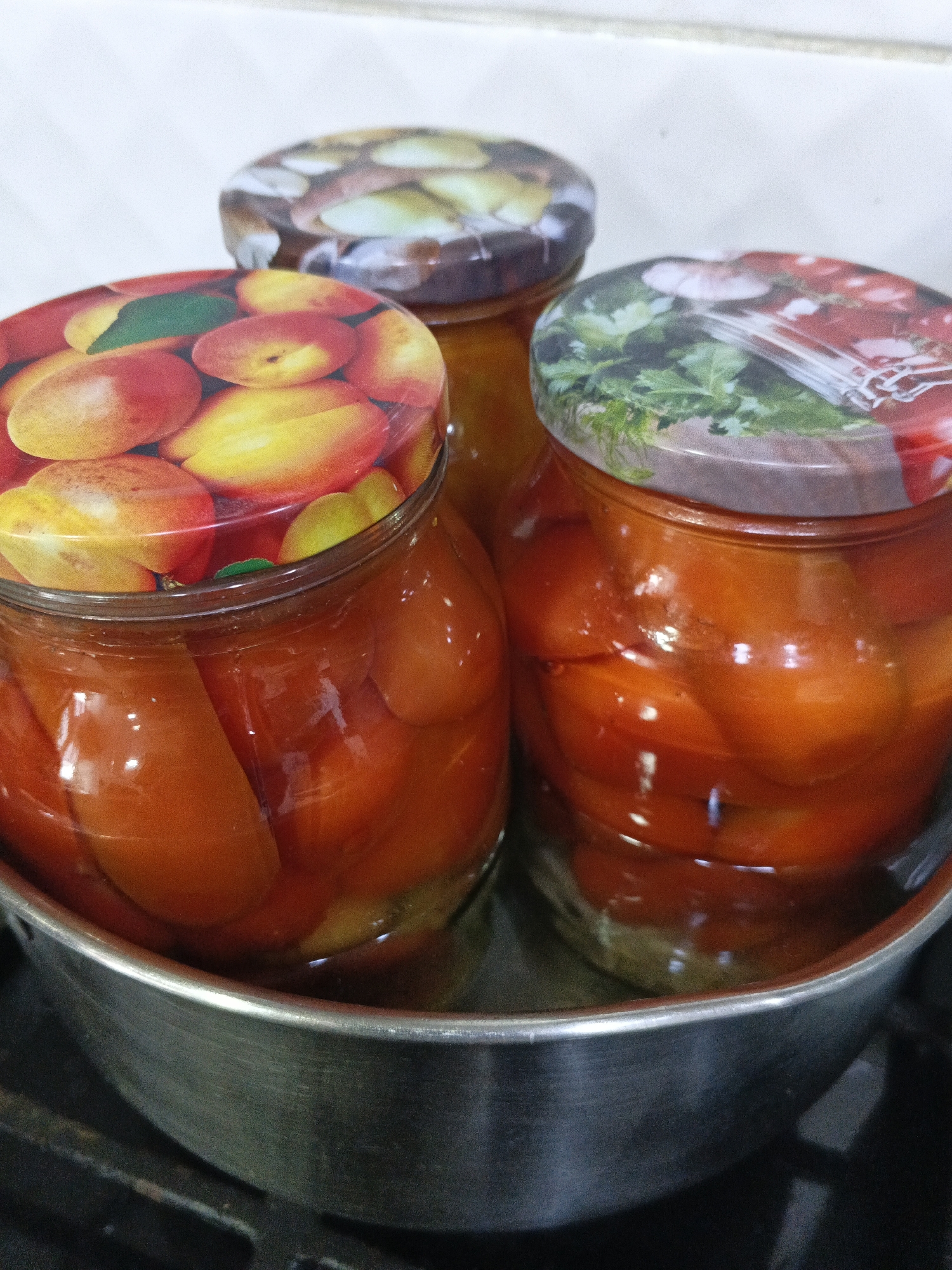 Three glass jars filled with preserved red bell peppers are arranged in a circular metal pan. The jars have decorative lids: one depicts sliced apricots, another shows fresh herbs, and the third is partially visible with a similar decorative pattern. The jars are placed on a stovetop. The scene is set in a kitchen in Medvedyovka, Russia.