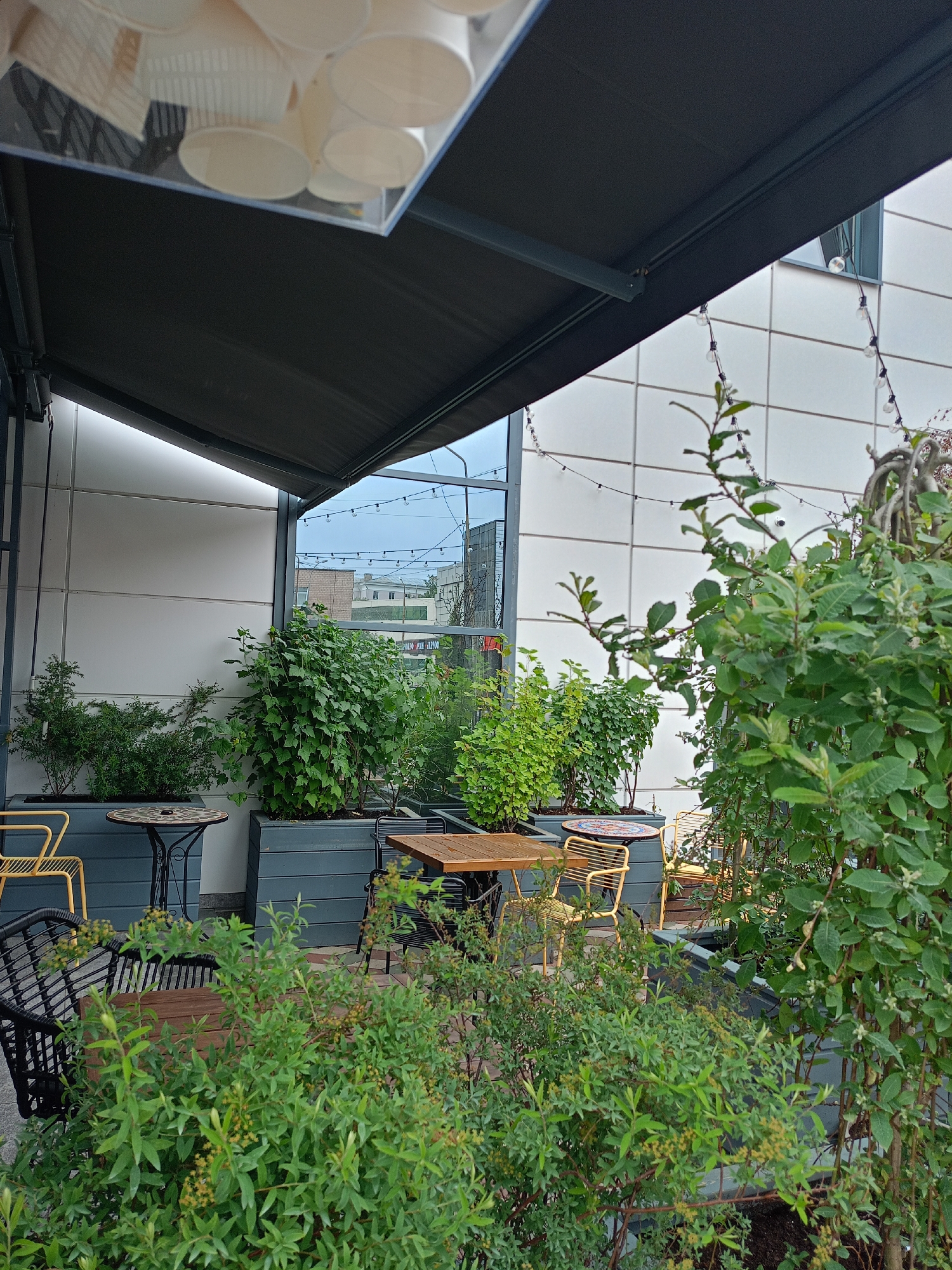 An outdoor cafe seating area is shown, with tables and chairs surrounded by planters filled with various green plants. A dark gray awning extends over the seating area. The background features a modern building with a white tiled facade and a large window. String lights are visible along the top of the building. The scene is set in an urban environment, possibly a cafe or restaurant patio.
