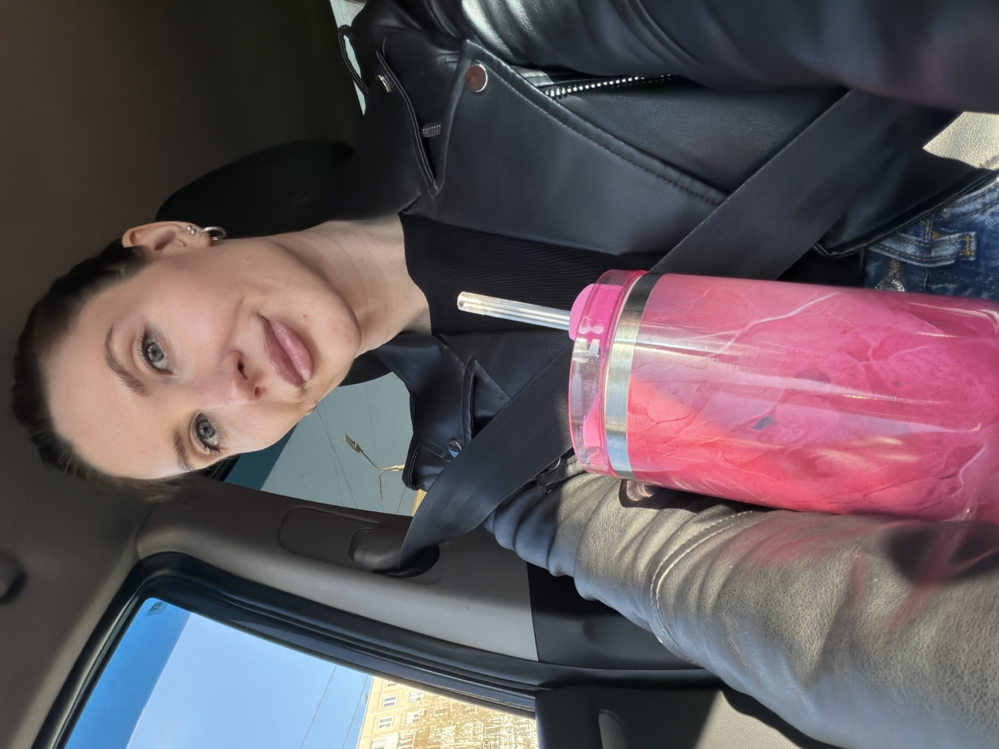 A person wearing a black leather jacket and jeans is inside a car. A large pink tumbler with a straw is in the car. A seatbelt is fastened over the person and the tumbler. Outside the car window, a building with multiple windows is visible. Based on the location and architectural style, it is inferred that the scene is set in Perm, Russia.