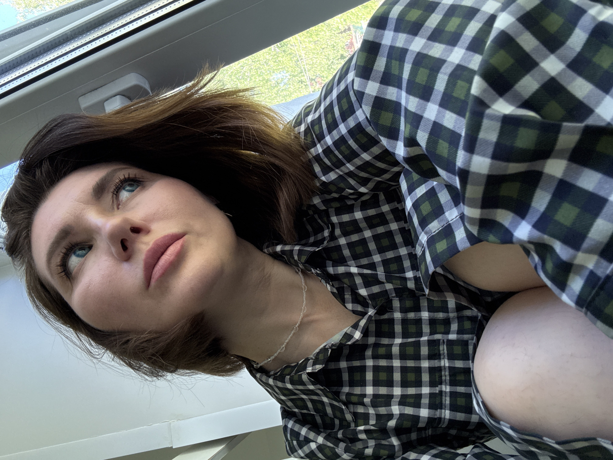 A low-angle shot shows a woman with brown hair and blue eyes, wearing a green and white plaid shirt. She is looking upwards. A window is visible in the background. There is no indication of the location being Chaikovsky, Russia.