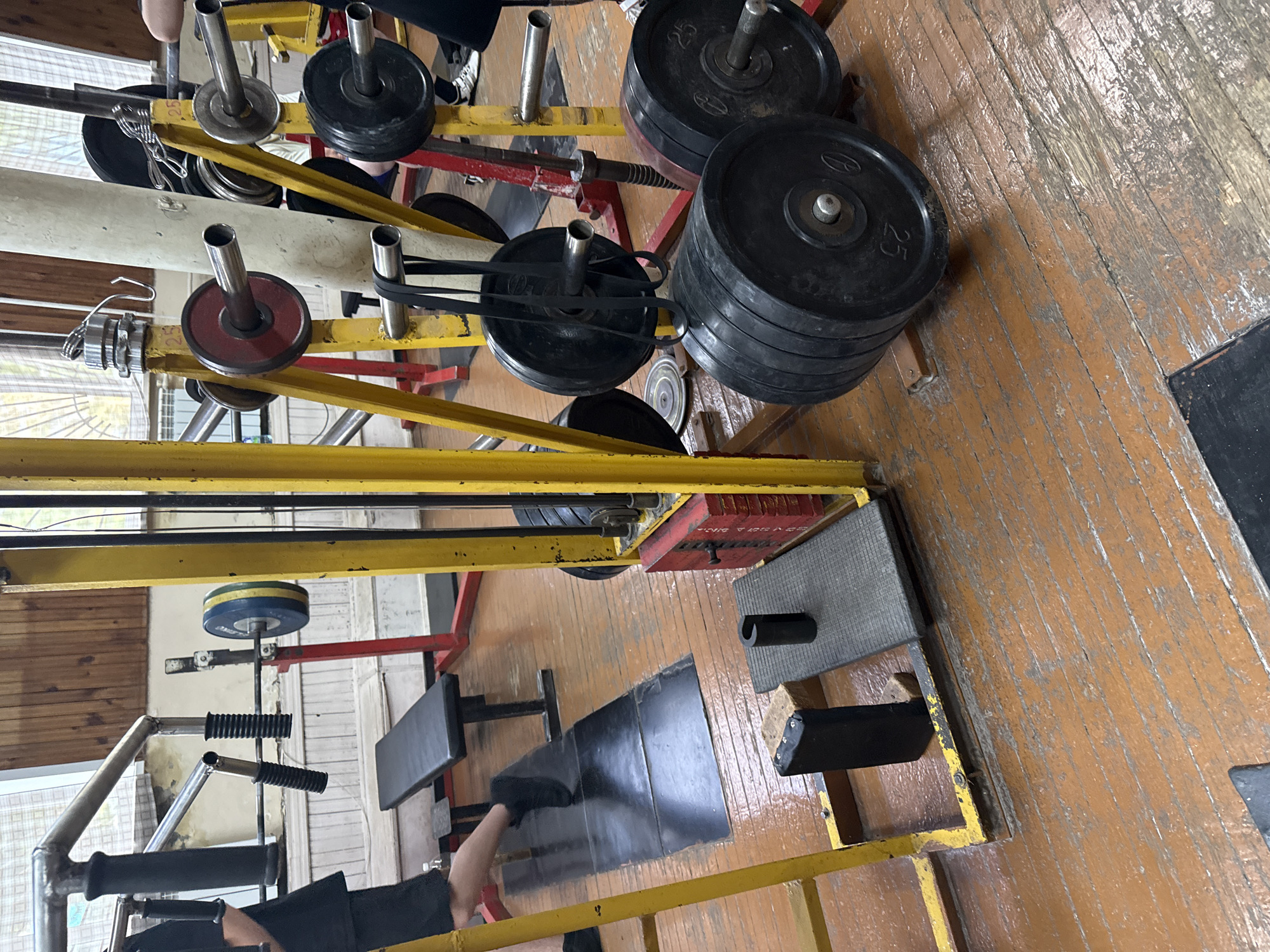 This is an indoor photograph taken at a gym in Kazan, Russia. The gym appears to be older, with worn wooden floors and dated equipment. In the foreground, a set of stacked weight plates, some marked with "25", are visible on a barbell. To the left, a black weight bench is positioned near a metal rack holding a barbell with weight plates. Further back, a person's legs are visible, wearing black shorts and athletic shoes, suggesting they are either using the equipment or moving through the gym. Other weight plates and gym equipment, including a yellow metal frame, are scattered throughout the scene. The overall impression is of a functional but somewhat unkempt training facility.
