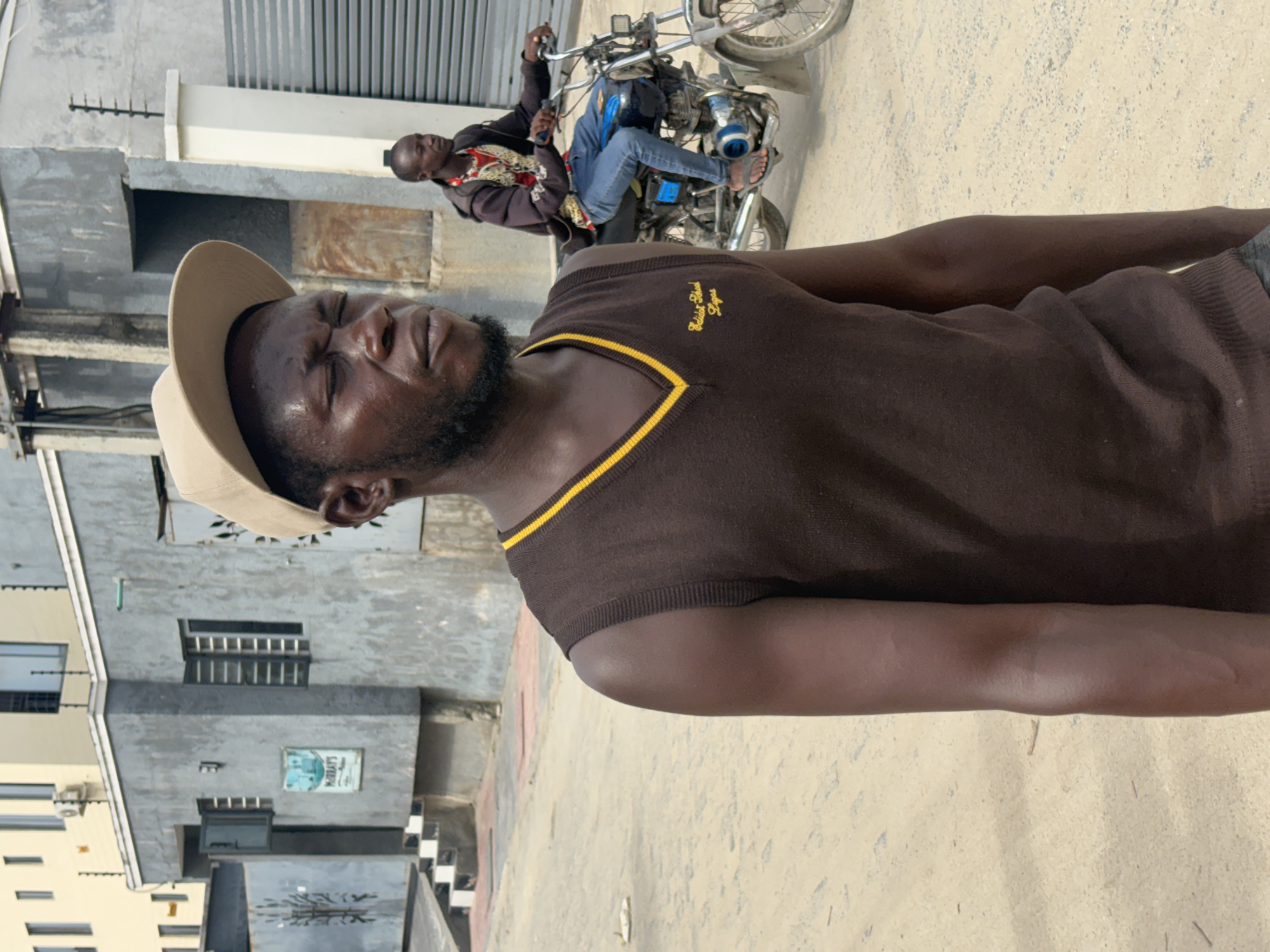 An outdoor scene in Igboefon, Nigeria, depicts two male individuals on a dusty unpaved road during daylight. In the foreground, a man of medium complexion stands slightly off-center to the left, facing right with his eyes closed or squinting. He wears a beige baseball cap, a dark brown V-neck sleeveless sweater with yellow trim, and "Eddidat School Lagos" embroidered on the left chest. He has a beard, and sweat is visible on his forehead. In the midground, to the right and slightly behind the foreground subject, another man of medium complexion operates a blue and silver motorcycle. He wears a dark hooded jacket over a red and white patterned shirt, blue jeans, and flip-flops, holding the handlebars. The motorcycle appears to be in motion, stirring dust. The background features various urban structures: a grey stucco-finished building with louvered windows and a "MURRAY'S" sign is visible to the left, behind a low wall with security spikes and a grey metal gate featuring a tree motif. A set of black and white steps leads up to this building. Further back, a lighter yellow multi-story building with windows and an air conditioning unit is partially visible. To the right, a grey roll-down gate and a white concrete pillar are present. Utility poles or concrete structures are also visible.