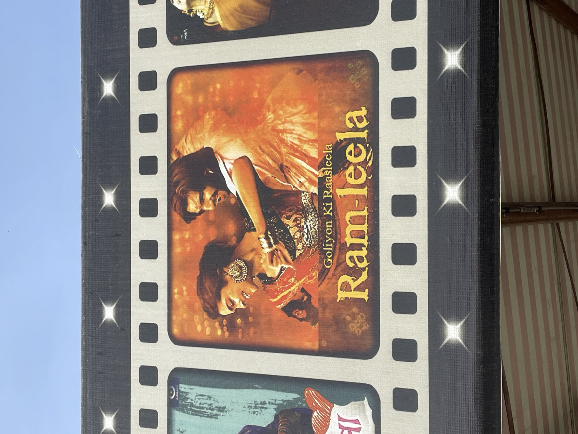 A vertically-oriented close-up shows a panel resembling a film strip with various movie posters. The central image is the poster for the Bollywood film "Goliyon Ki Raasleela Ram-Leela," featuring a stylized image of a couple. The background of the poster is an orangish hue. The overall panel has a dark gray background with silver or white film strip borders. The scene appears to be outdoors, as evidenced by the blue sky visible in the upper left corner and the striped canopy visible in the background. Based on the location information provided, the scene is in Surmaiguda, India.
