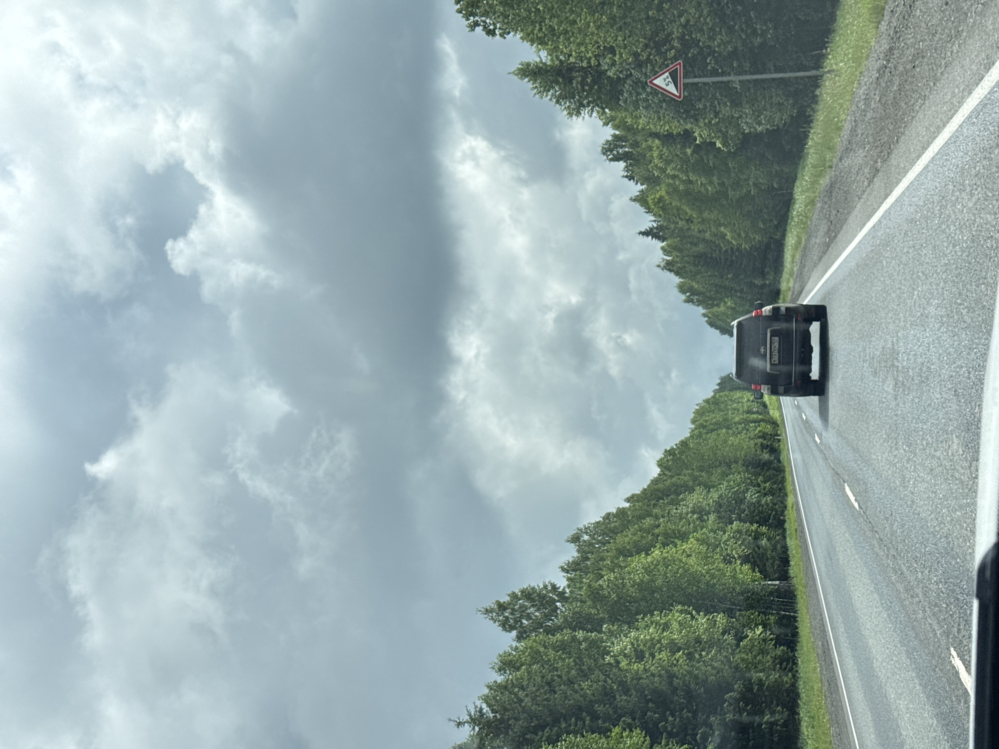 The image captures a view from a vehicle driving on an asphalt road in what appears to be a rural or forested area, possibly near Anninsk, Russia, during daytime under an overcast sky. The perspective is looking forward, following a black SUV or van.

The road ahead is wet or damp, suggesting recent rain, with visible white lane markings. Dense green trees line both sides of the road, forming a continuous forest canopy alongside the route. The sky above is largely covered by heavy, grey and white clouds, indicating cloudy weather.

A triangular warning sign with a red border is prominent on the right side of the road, set against the trees. The sign features a black arrow pointing upwards with "5%" written inside, indicating a 5% uphill gradient. The black SUV or van in front has a visible Russian license plate, which reads "A 120 ХО 99 RUS". The "99" in the license plate corresponds to the region code for Moscow, Russia. No people are discernible in the image.