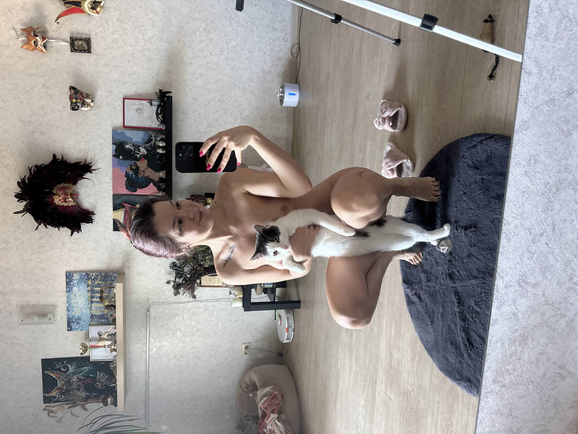 A female individual is depicted in a residential interior, taking a mirror selfie while holding a bi-colored cat. The woman, appearing nude, is squatting on a dark gray circular rug. She has fair skin, dark hair with reddish highlights, and a text tattoo on her upper left torso. She holds a black smartphone with three rear camera lenses in her right hand; her fingernails are painted bright pink, and she exhibits a subtle smile.
She cradles a white cat with black markings on its head, back, and tail. The cat, possessing yellowish-green eyes, stands upright on its hind legs, supported by the woman's left arm and lap, facing the camera.
The interior walls are light gray with a textured finish. On the upper left wall, a light-colored floating shelf displays a framed painting of a cat-like figure in a suit, a golden trophy cup, various small figurines, and a nocturnal cityscape painting. A simple frosted glass wall sconce is mounted above the shelf. To the right, a large ornate mask composed of black and red feathers with gold accents hangs on the wall. Further right, a black floating shelf is positioned below several decorative masks, including a fox-like mask, a patterned harlequin mask, and a jester's hat/mask. The black shelf holds framed artwork, including a depiction of a woman with a blue mask kissing Batman, another framed sketch, a black feathered mask, and small white figurines.
The floor is light wood-grain laminate. In the far left background, a beige beanbag chair contains folded light-colored fabric. A white robot vacuum cleaner is visible on the floor near European-style electrical outlets. A black square end table is positioned in the mid-left background, topped with candles and other decorative items. On the right side of the frame, a white and blue pet water fountain is on the floor, connected to a wall outlet. A silver and black camera tripod stands on the far right. Two pink, bow-adorned slippers and a small brown mouse-shaped cat toy are on the floor, partially reflected in the mirror.