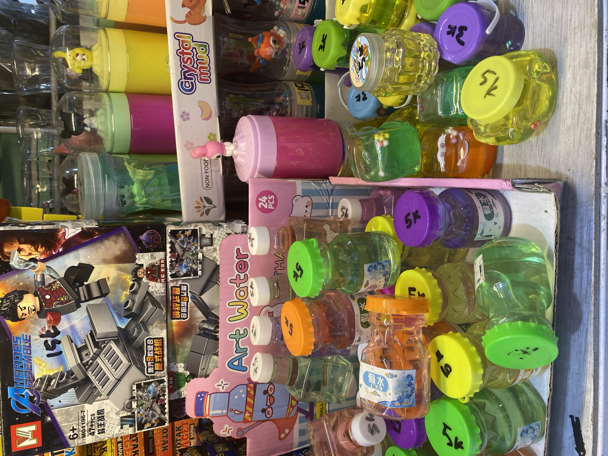 This image depicts a stall selling toys in Lamteumen Barat, Indonesia. The stall is filled with various colorful plastic toys, including action figures, slime containers, and novelty items. In the foreground, there are several small, clear plastic bottles filled with colorful liquids and some with tiny decorative elements, likely intended for children. These bottles have brightly colored caps, some marked with what appear to be prices or identifiers. Behind these, larger containers of slime and other novelty toys are visible, some with cartoon character designs. The overall impression is a vibrant and busy market stall offering a wide assortment of children's toys.