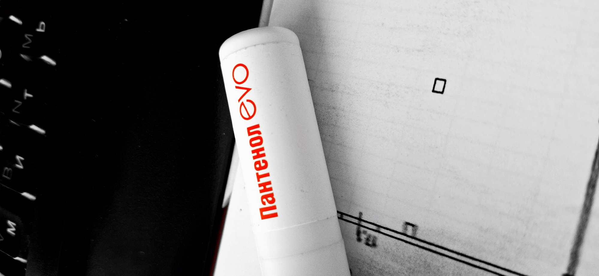 The image, captured in Chaikovsky, Russia, presents a close-up view of a white cylindrical object bearing red Cyrillic text "Пантенол evo" (Pantenol evo). The object lies horizontally across a white sheet of paper that exhibits printed black lines and a small black square. A dark-colored computer keyboard, partially visible in the upper background, displays keys with faint letters including 'M', 'B', 'И', 'N', 'T', 'М', and 'Ь'. The scene appears to be an indoor setting, likely a desktop, with selective coloration emphasizing the red text against an otherwise desaturated black and white palette. No people or specific actions are visible.
