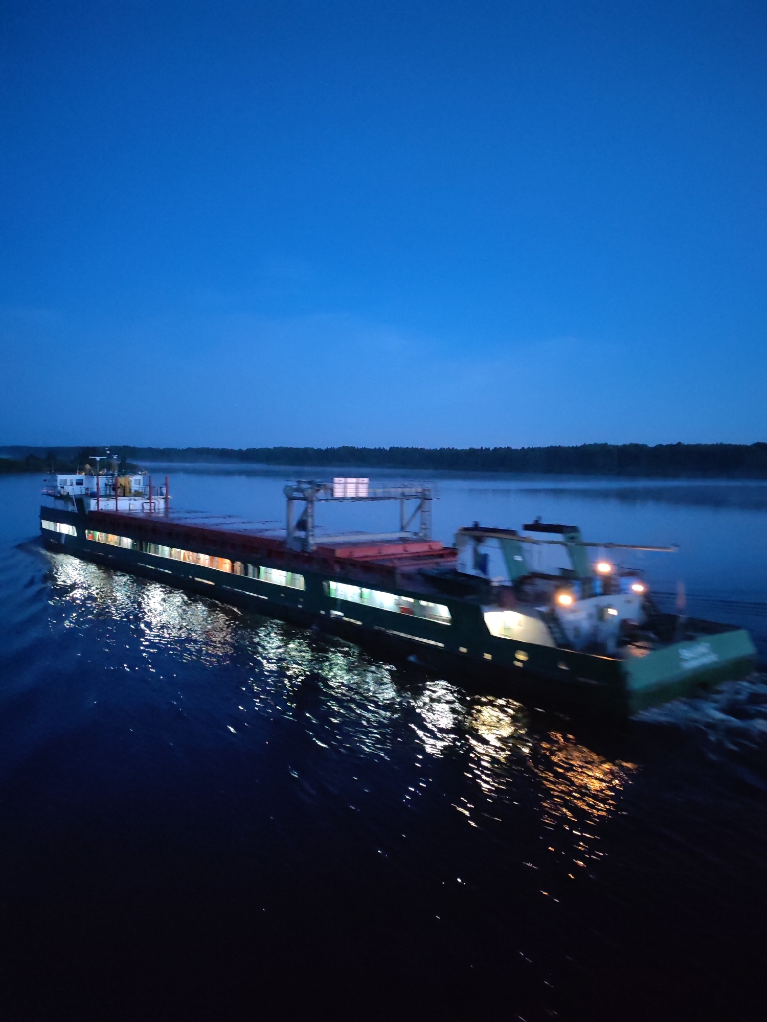A long, illuminated cargo barge moves on a body of water under a darkening sky. The barge has multiple light sources visible along its length. Reflections of these lights streak across the water's surface. A line of trees is visible on the horizon. The city of Gari, Russia, is not explicitly visible in the image, but the presence of such a large barge suggests the location is near a major waterway in the region.
