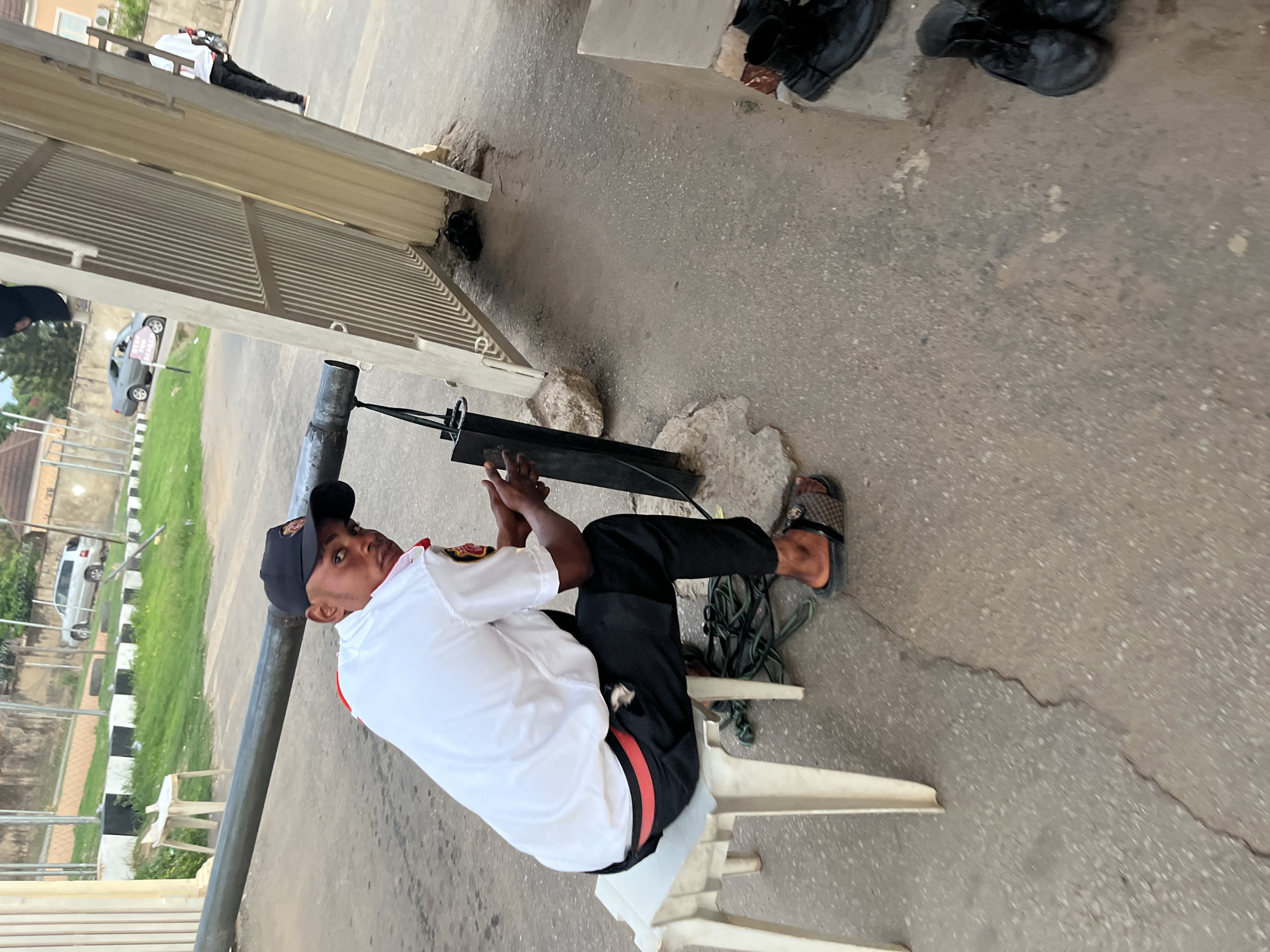 A security guard is seated on a white plastic chair next to a metal gate in Abuja, Nigeria. He is wearing a white shirt, black pants with a red stripe, a black cap, and slides. He is holding a black device. Nearby are work shoes and some coiled green electrical cords. Behind the gate is a glimpse of parked cars and greenery. The ground is concrete.