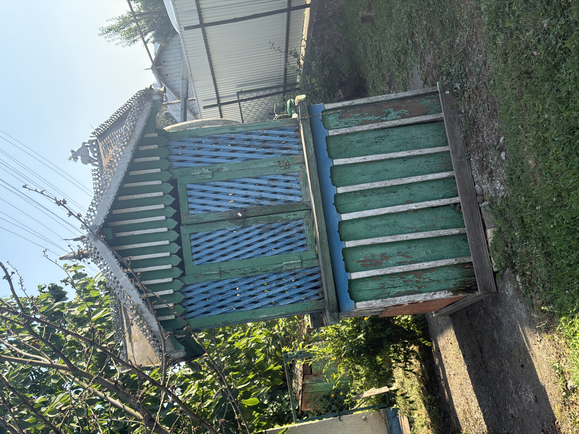 The image shows an old, weathered, two-tiered wooden structure with a decorative, peaked roof, painted green with blue lattice-work doors. The structure is situated in a yard with grass and dirt pathways. A tree with green leaves is to the left, and a metal fence is in the background. The overall scene suggests a rural or suburban outdoor setting.