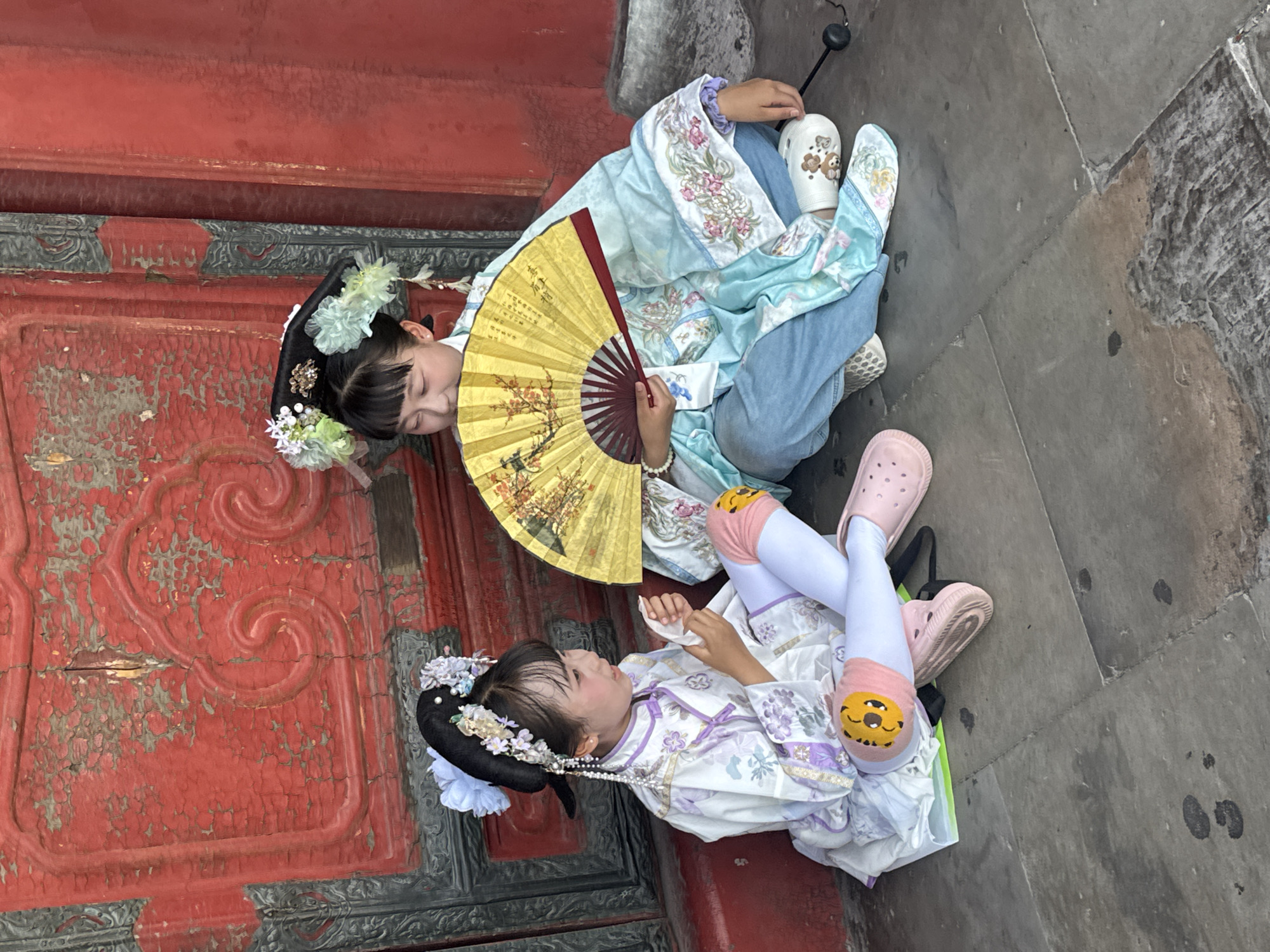 In Dongcheng District, China, two young girls are seated on a stone ground in front of a weathered red door. The girl on the left, wearing a white and purple traditional Chinese outfit with floral embroidery, holds a white tissue in her hands. Her legs are crossed, and she wears white stockings with pink shoe-like coverings featuring a cartoon character. The girl on the right, dressed in a light blue traditional Chinese garment over blue jeans, holds open a large yellow fan adorned with intricate artwork. Both girls have black hair styled with traditional Chinese hair ornaments featuring flowers and pearls. The red door behind them exhibits peeling paint and ornate carved details. The scene suggests a cultural outing or reenactment.