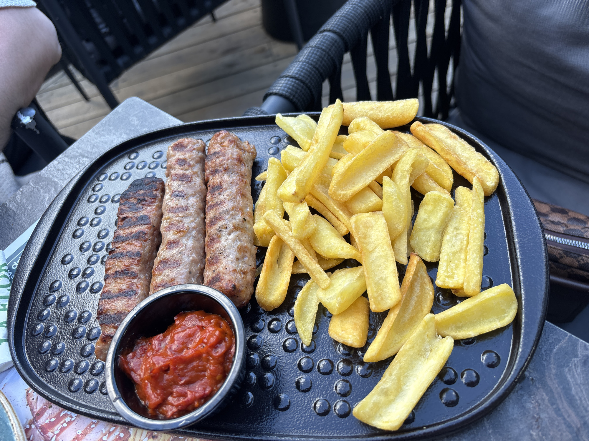 The image shows a close-up, high-angle view of a meal served on a black, textured platter. The meal consists of four grilled sausages and a serving of french fries, accompanied by a small metal bowl filled with a red relish or sauce. The sausages appear to be seasoned and have distinct grill marks. The french fries are cut into thick wedges and have a golden-brown hue. The platter is placed on a table with a wooden surface, and a dark woven chair is visible in the background. A person's arm, wearing a white bracelet, is also visible in the upper right corner. The overall setting suggests an outdoor dining experience, possibly at a restaurant in the city of Nesebar, Bulgaria, given the context provided. The image is well-lit, with natural light illuminating the food and its surroundings.