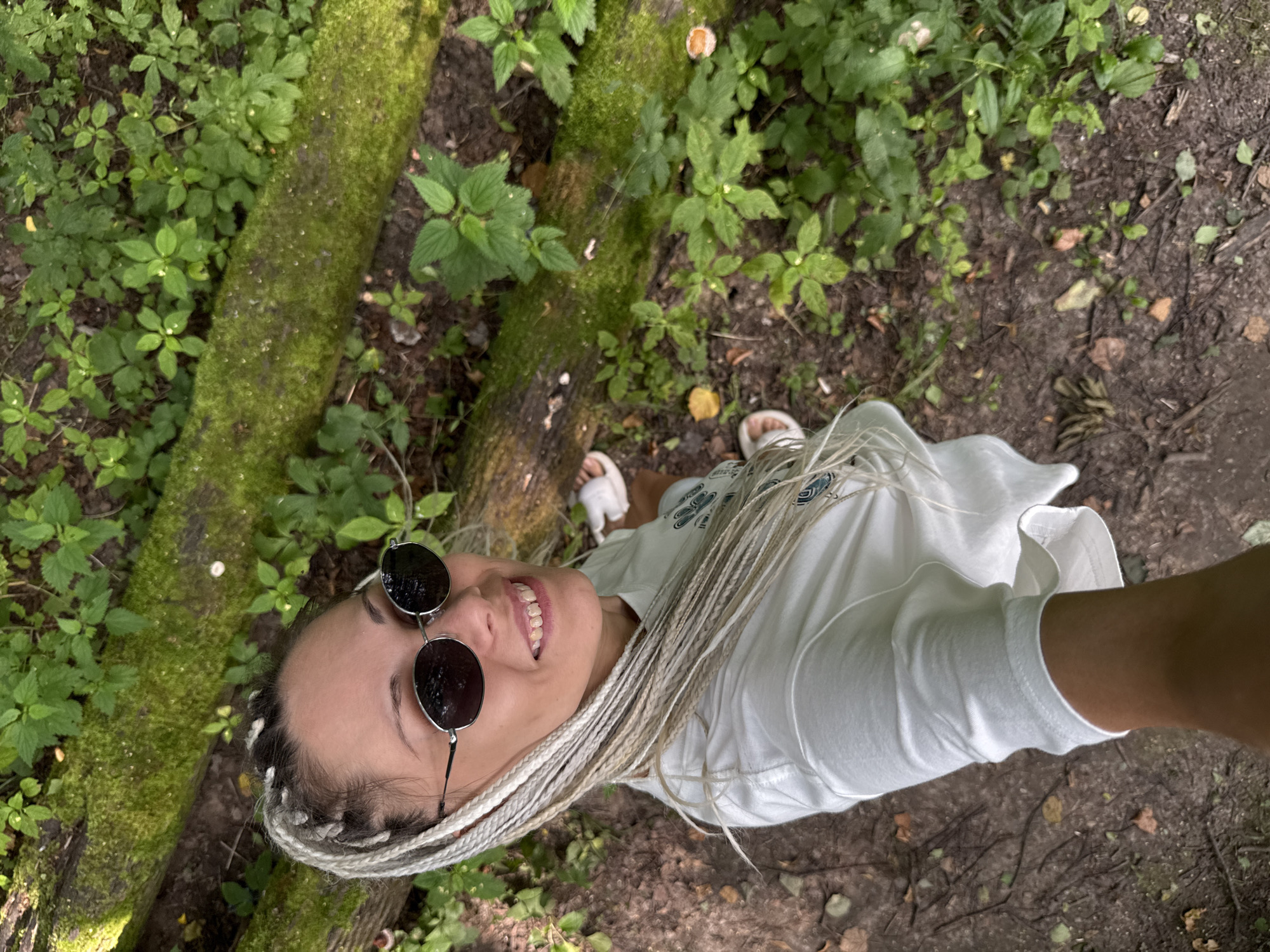 A woman with blonde braids and sunglasses is taking a selfie in a wooded area. She is wearing a white t-shirt and white sandals. The ground is covered with dirt, leaves, and mossy logs. She is smiling at the camera. The location is identified as Rusanovo, Russia.