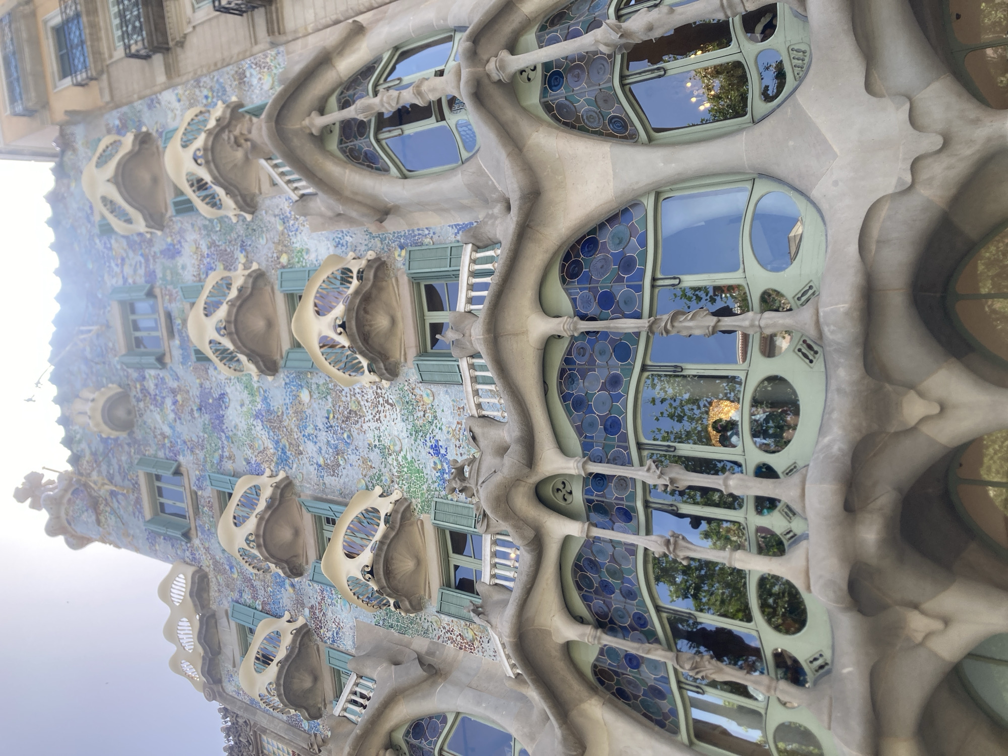 The image showcases the distinctive architecture of Casa Batlló in Barcelona, Spain. The building's facade is adorned with a vibrant mosaic of blue, green, and white tiles, interspersed with organic, bone-like balconies. Several windows are visible, featuring irregular shapes and decorative stained glass in shades of blue. The building's facade itself has a fluid, undulating quality, reminiscent of natural forms.
The image appears to have been taken during the day, under clear skies, with bright sunlight casting reflections on the windows. The overall impression is one of intricate design and artistic innovation, typical of Antoni Gaudí's work. There are no people or specific activities depicted, the focus is solely on the architectural details of the building. The image is a close-up view, emphasizing the texture and patterns of the facade and the unique design of the windows and balconies. No visible text is present in the image.