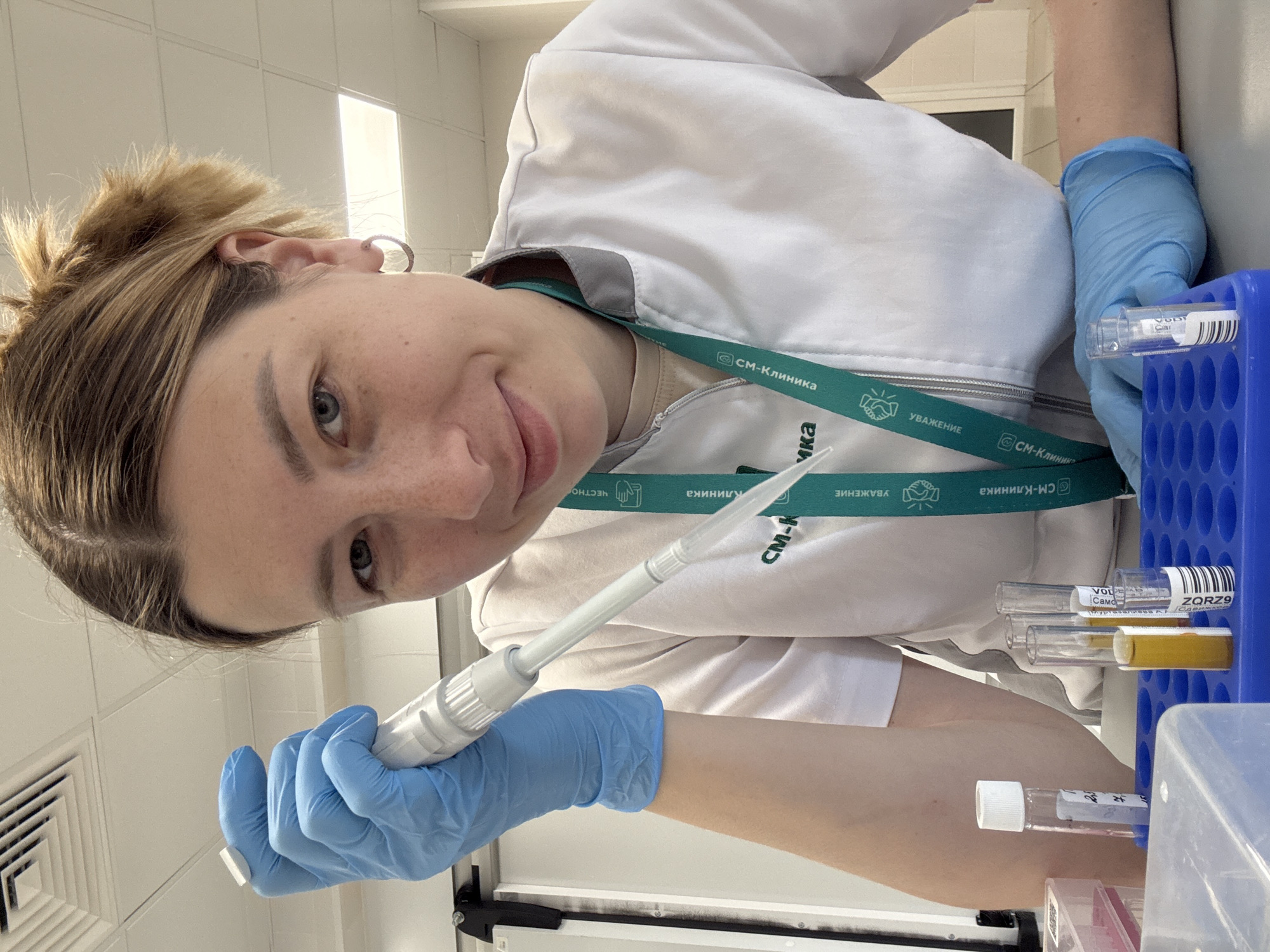 A young woman wearing a white lab coat and blue gloves is working in a laboratory, specifically in Moscow, Russia, as indicated by the "CM-Clinic" label on her lanyard. She is using a pipette to transfer liquids from a test tube rack containing multiple tubes, some with yellow liquid. The background includes laboratory equipment and ventilation.