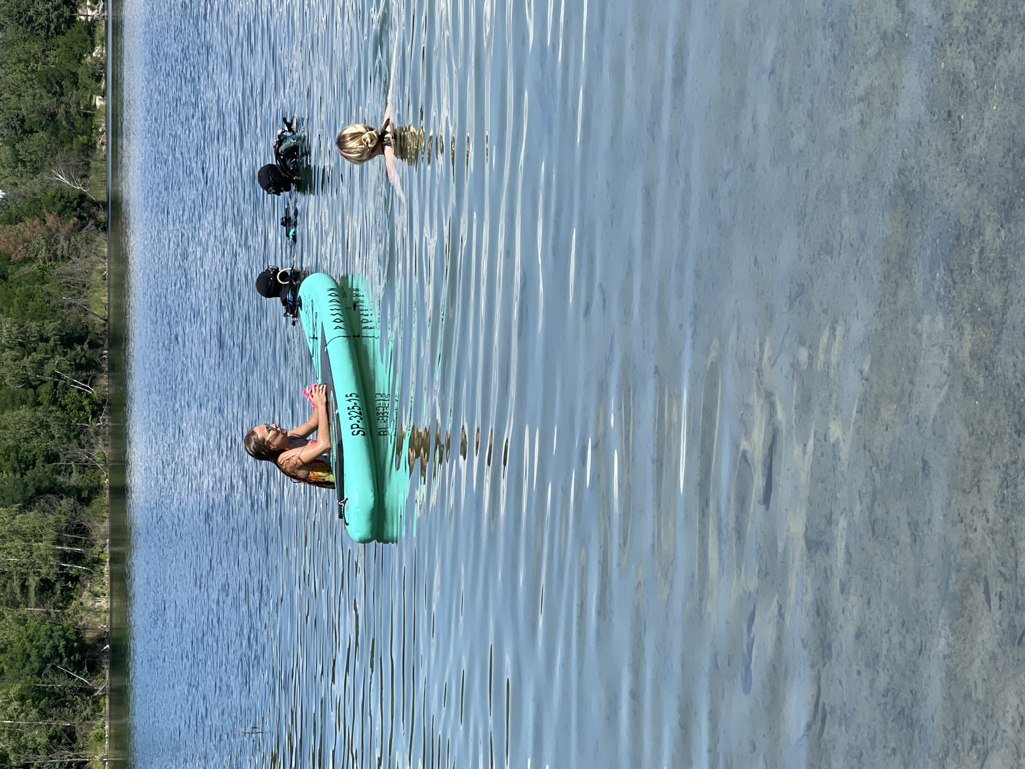 The image shows a body of water, likely a lake, with a clear, shallow bottom visible in the foreground. Trees and vegetation line the distant shore. In the water, there are several individuals and an inflatable raft.
On the raft, a young woman wearing a colorful swimsuit is seated, holding a pink object. The raft is teal and has text visible, including "SP-325-15" and what appears to be a brand name that starts with "POSSIDION."
Further out in the water, two individuals in full scuba gear are present, with their heads and shoulders above the surface. They appear to be in shallow water.
In the middle ground, a woman with blonde hair pulled back in a bun is swimming, facing away from the camera. The water is calm with gentle ripples. The lighting suggests it is daytime, with sunlight reflecting off the water's surface. The scene appears to be a recreational area. There is no indication of the location being in the city of Korosten, Ukraine.