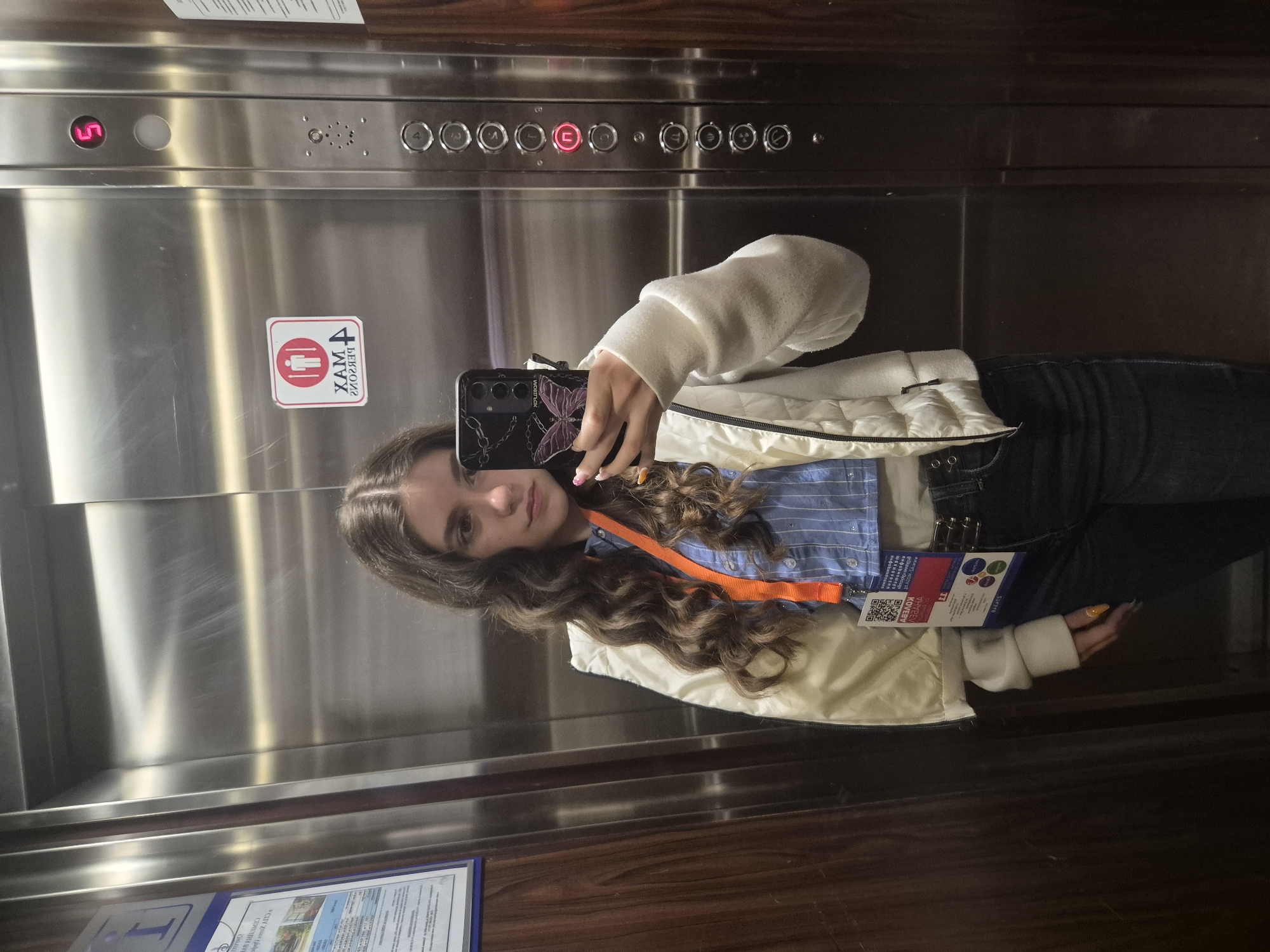 A young woman is taking a selfie in an elevator. The elevator has stainless steel doors and wood paneling on the side walls. The elevator control panel indicates that the elevator is currently on the 5th floor. A sticker in the elevator indicates a maximum capacity of 4 persons. The woman is wearing a white jacket over a blue striped shirt, with dark-colored jeans. She has an orange lanyard around her neck with an ID badge. The ID badge has the text "Коледух" on it and a QR code. A sign is visible in the lower-left corner of the elevator, presumably related to information about Devin, Bulgaria, which is the location. The elevator is indoors.