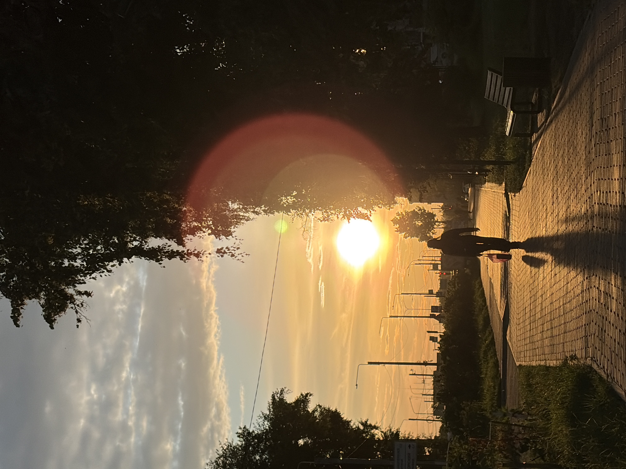 A person walks down a brick pathway in Chaikovsky, Russia, during sunset. The sky is a gradient of orange and blue, with wispy clouds. The sun is a bright orb, casting long shadows from the trees and the person. A red lens flare is visible on the right side of the image. On the right side of the pathway, a wooden bench and trash receptacle are situated. In the background, streetlights and buildings can be seen. The overall scene is serene and picturesque.