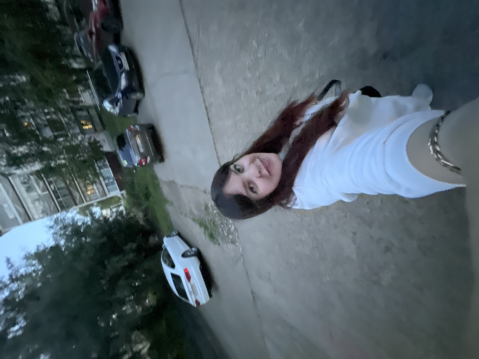 A female individual is captured in a selfie at dusk on a paved residential street in Noviy, Russia. The woman, positioned in the lower right, has dark, shoulder-length hair and is smiling directly at the camera. She wears a white long-sleeved top, with a metallic chain-link bracelet visible on her right wrist as she holds the camera.
The paved street, showing signs of wear with cracks and scattered debris, extends into the background. A white two-door sedan, consistent with an Audi A5 Sportback, is parked facing away from the camera on the left side of the street. On the right, further back, a dark blue sedan is parked, followed by a red sedan. Beyond the vehicles, large, dark green trees line the street.
In the background, multi-story apartment buildings are visible. One prominent light-colored building on the right features multiple windows and balconies, with interior lights illuminated in some units. The sky overhead is a light blue-grey, indicative of late evening or dusk conditions.