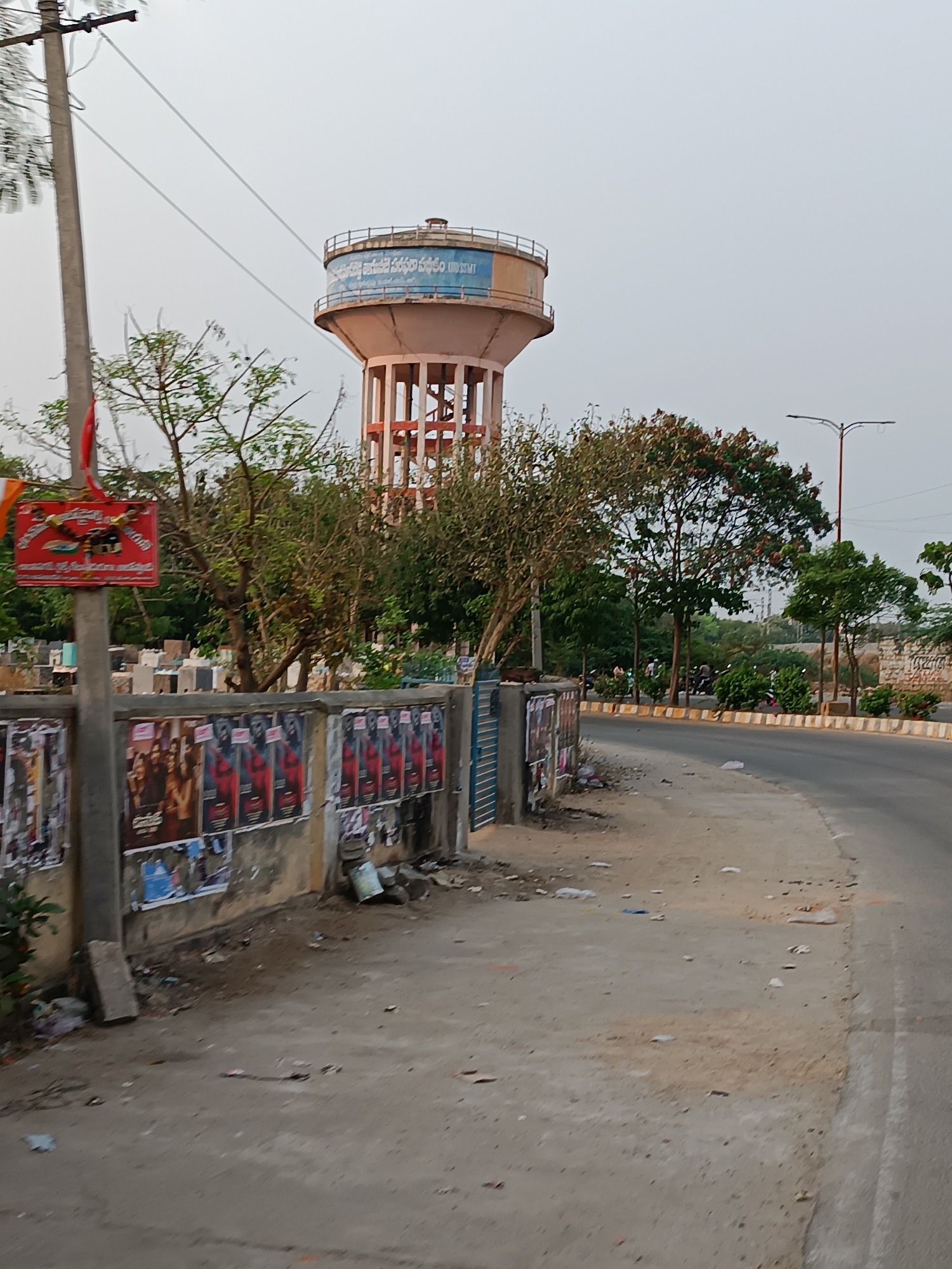 The image shows a street scene in Dharmavaram, India. A tall, light pink water tower with a blue band containing writing in Telugu dominates the background. In the midground, a low, weathered concrete wall, covered with torn and peeling movie posters, lines the street. A red sign is affixed to a utility pole. A graveyard appears behind the wall. A paved road curves to the right in the foreground and midground. Some litter is scattered on the ground between the wall and the road. There are some trees and light posts. People are visible on the road, walking and on motorbikes.