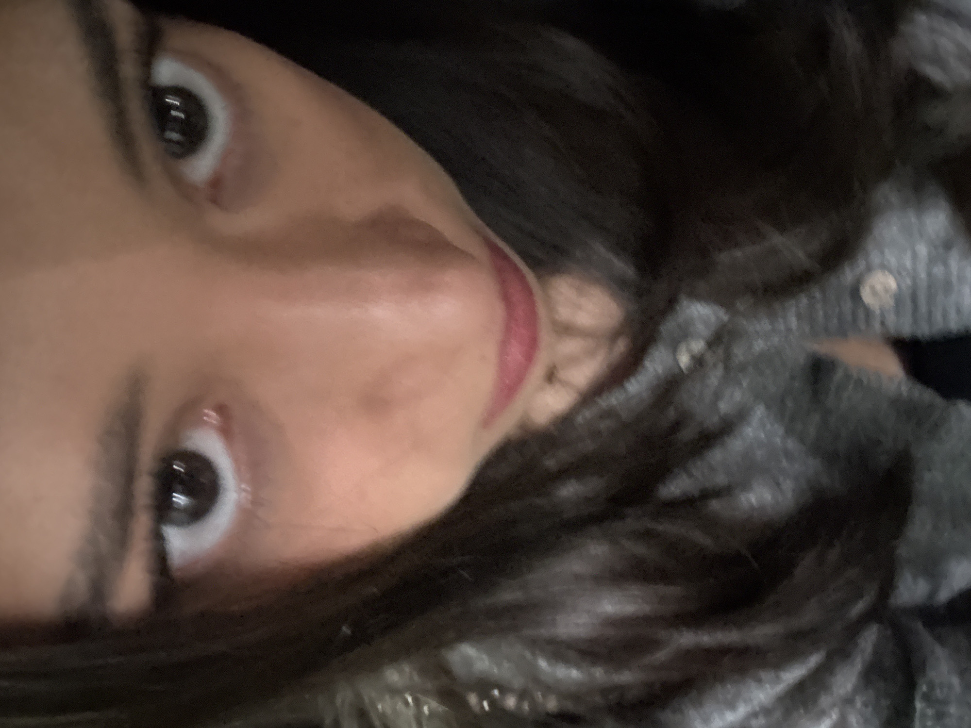 A close-up, slightly tilted selfie of a woman's face. She has dark hair, light skin, and dark eyes. She is wearing red lipstick and a gray sweater. The location is not discernible from the image.