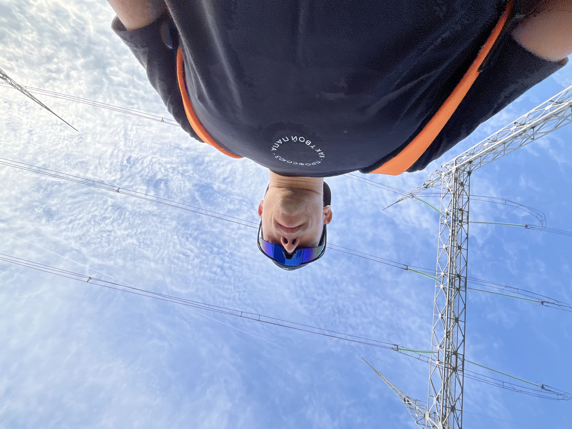 A low-angle selfie taken in what appears to be Chaikovsky, Russia, shows a man looking down at the camera, wearing sunglasses and a black t-shirt with orange accents. The background features a blue sky with patchy clouds and a metal power line tower with high-voltage wires. The text on the t-shirt is in Russian.