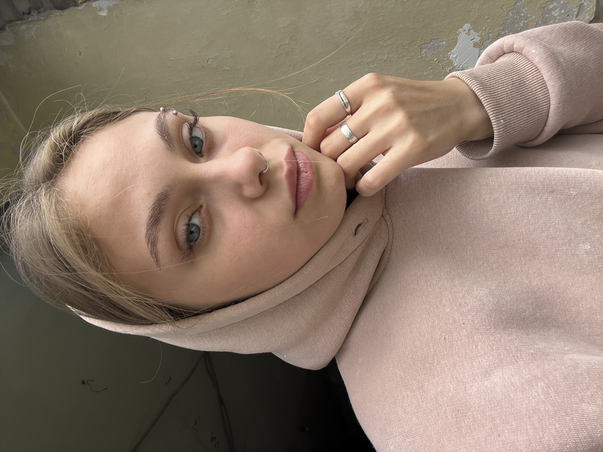 A close-up, slightly angled portrait shows a young woman with fair skin, blonde hair, and blue-green eyes. She is wearing a pink hooded sweatshirt, with the hood partially framing her face. The woman has a piercing on her eyebrow and a ring in her nose. Her hand, adorned with two silver rings, is near her face, partially obscuring her cheek. The background is a mottled combination of grey and beige, suggesting a weathered wall or surface. It is not possible to determine the location in the city of Фоки, Russia, from the provided image.