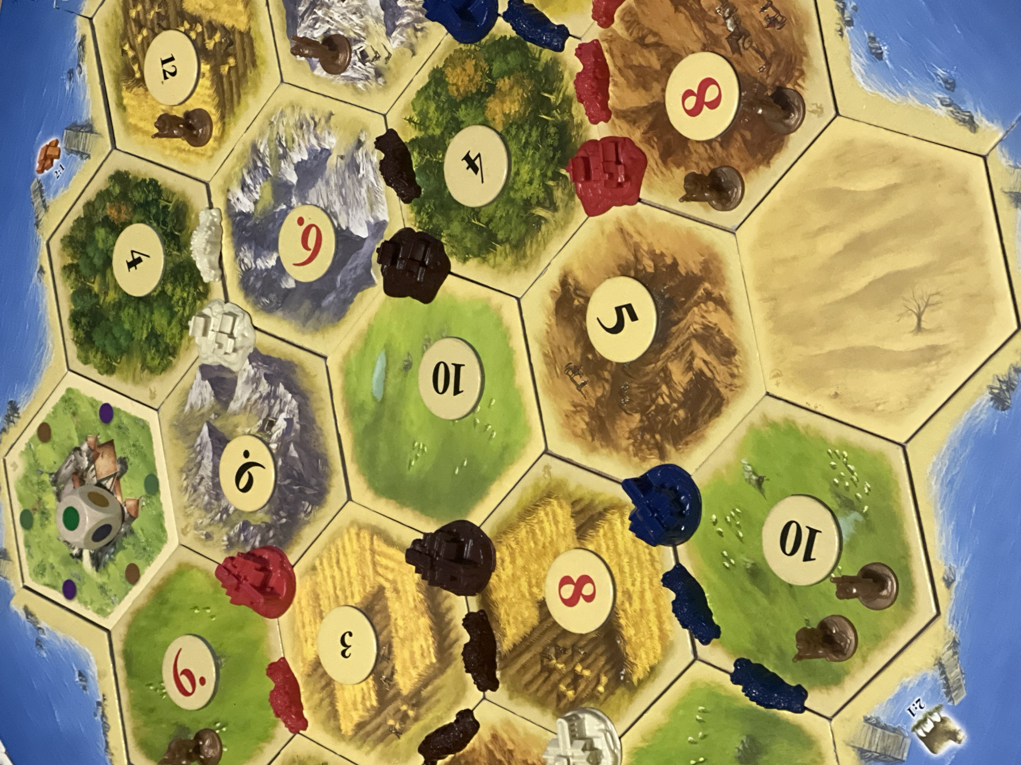 This is a close-up, high-angle photograph of a Catan board game. The game is set up on a flat surface, likely a table. The board is composed of hexagonal tiles representing different terrain types such as fields, forests, mountains, pastures, and a desert. Each tile has a number token, and many have game pieces on them.
There are several game pieces visible:
- Red settlements and roads are placed on the board.
- Brown wooden pieces representing settlers and possibly cities are also visible.
- Blue pieces, which could be roads or settlements, are present.
- White pieces, likely settlements or cities, are also scattered across the board.
- A six-sided die with green, white, and grey faces is resting on a green pasture tile.
- Several number tokens with red or black numbers are clearly visible: 3, 4, 5, 6, 8, 9, 10, 12.
- One tile depicts a port with a "2:1" marking.
- The background shows the blue sea surrounding the island.
The image does not contain any information that places it in the city of Varna, Bulgaria.