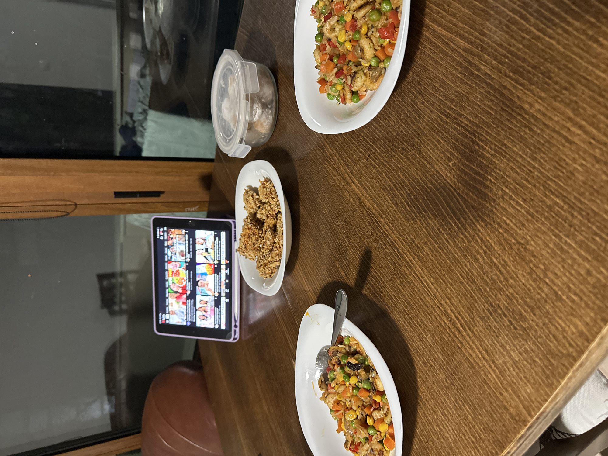 A tabletop is set with several dishes of food, including one plate with fried rice and a spoon, another plate of what looks like fried chicken, and a food container containing cookies or biscuits. A tablet displays content on the side, resting adjacent to a leather-like item. The tabletop has a wooden grain and seems to be indoor due to the proximity of the window, and it's not possible to determine any more about its location than that.