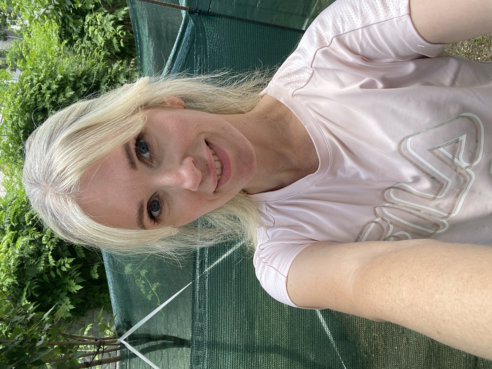 A Caucasian woman with blonde hair and blue eyes is pictured from the chest up, taking a selfie outdoors. She is wearing a pale pink t-shirt with a large, reflective "Fila" logo on the front. The background is blurred, showing green foliage and a green mesh fence. The lighting suggests it is daytime.
