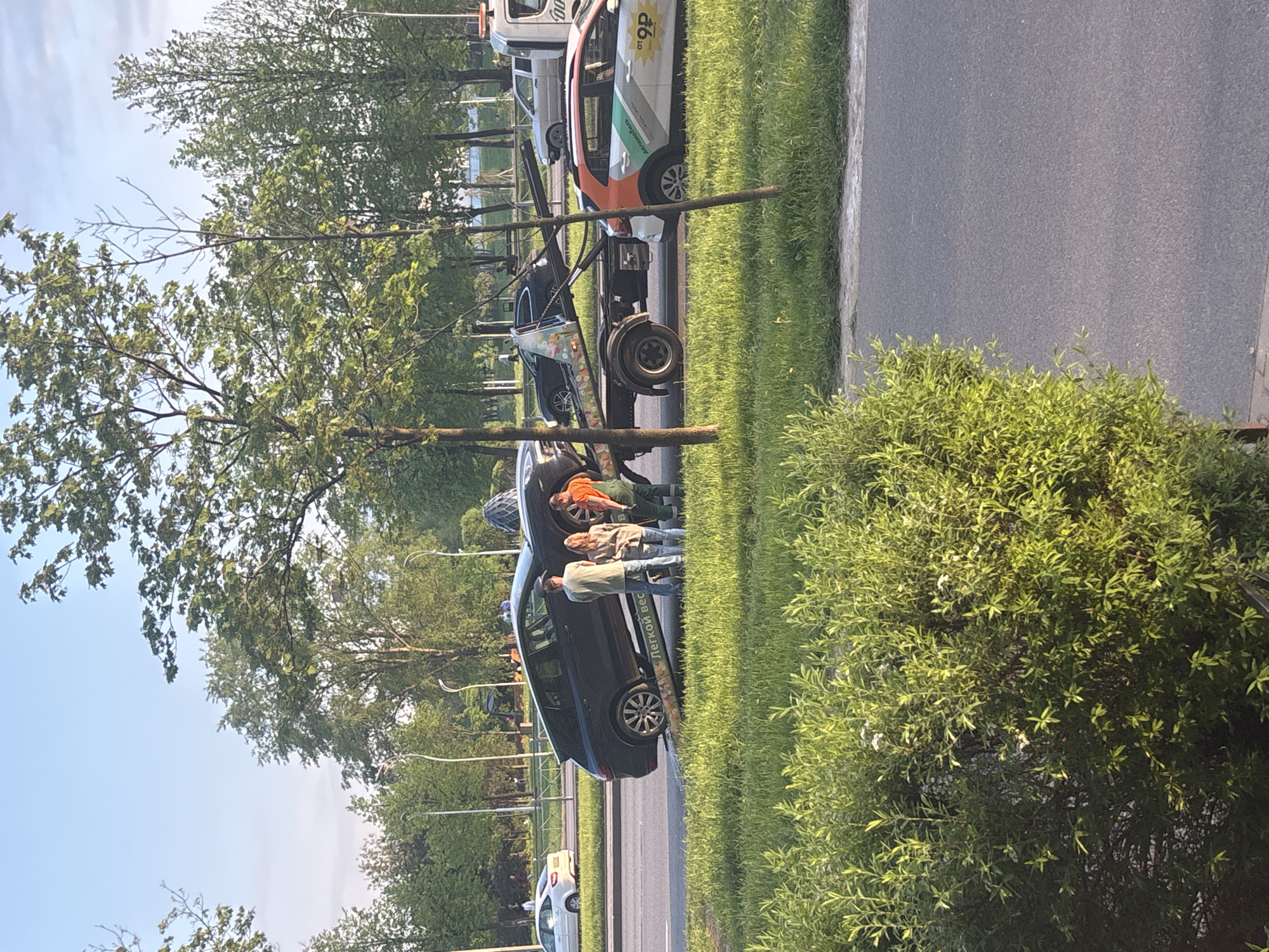 The image shows a street scene in Unknown, Unknown. A dark-colored car is parked on the grass verge next to the road. Three people are standing near the car; one is wearing an orange top, another a tan jacket, and the third a green shirt. A tow truck is parked further up the road. There are trees lining the road, and a building is visible in the background. The sky is blue. The ground appears to be covered with grass and foliage.