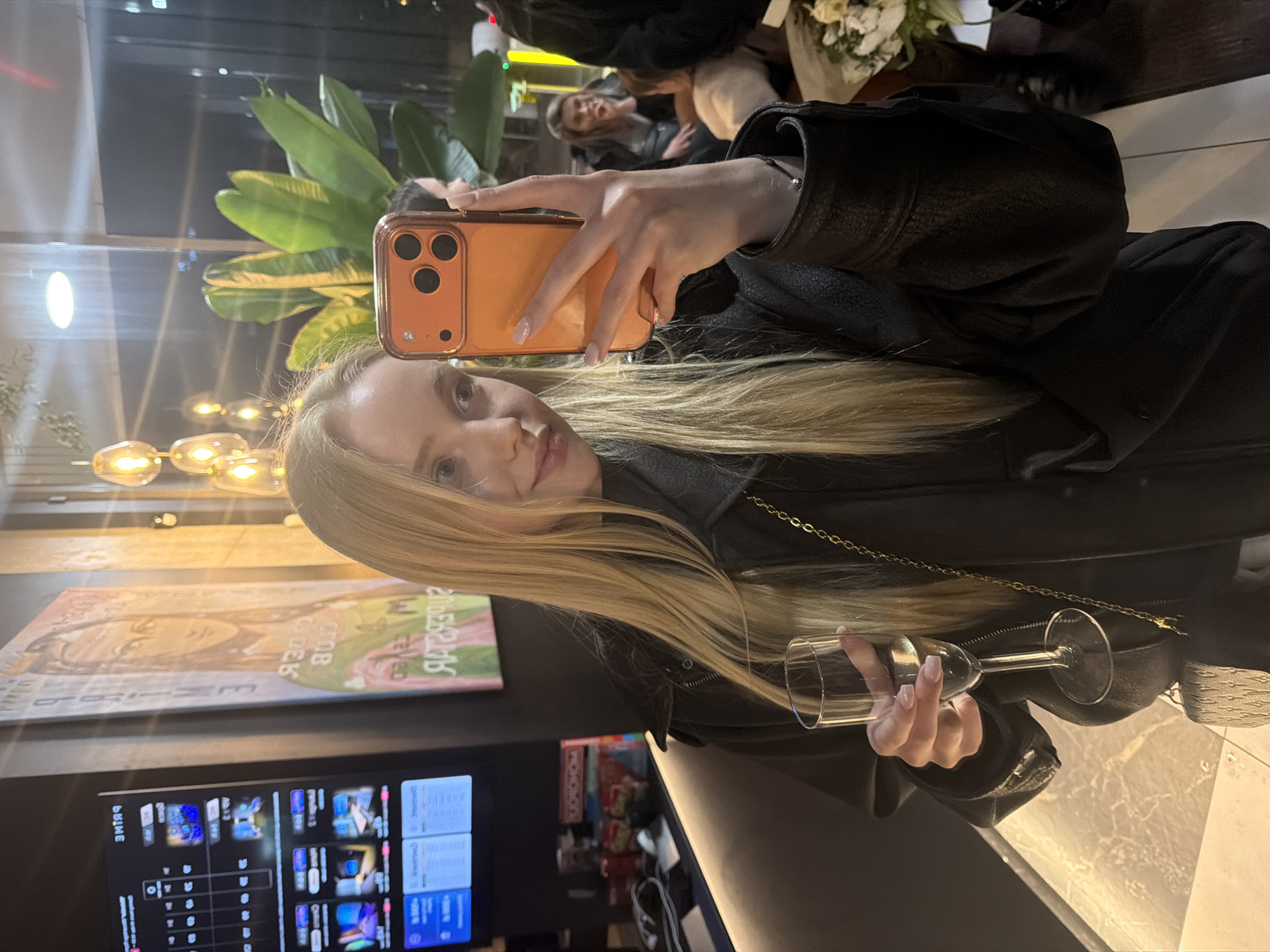 A blonde woman holds a smartphone in her right hand to take a selfie, while a wine glass is held in her left. She is wearing a black leather jacket and has a gold chain necklace. She is indoors in a modern setting with a large television screen displaying menus. A piece of artwork hangs on the wall above the screen. There are decorative hanging lights and a potted plant in the background. Other individuals are visible in the background. The location is the city of Unknown, Unknown.