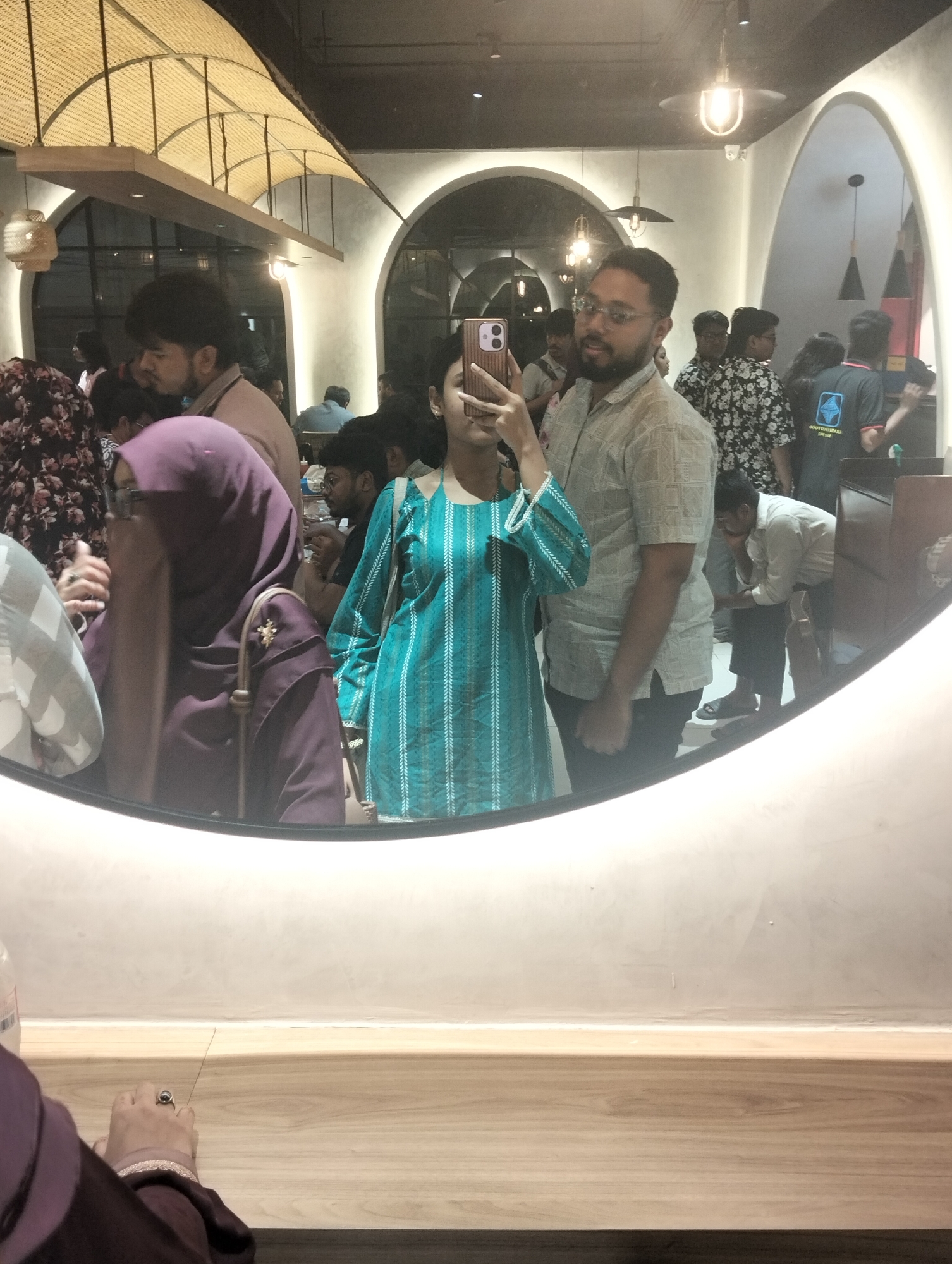 A mirror selfie reflects an indoor scene filled with people. The selfie is taken by a person wearing a turquoise and white patterned top, holding a tan-colored smartphone. Behind this person stands a man in a light-colored button-down shirt and glasses. The mirror's reflection captures a crowded room with various individuals, some standing, others seated, and others moving about. At least one person in the crowd is wearing a floral-patterned shirt. One person is wearing a purple hijab. The background reveals architectural details, including arched doorways and a ceiling with woven light fixtures. The lower portion of the mirror frame rests on a wooden surface. Dhaka, Bangladesh is a probable location.