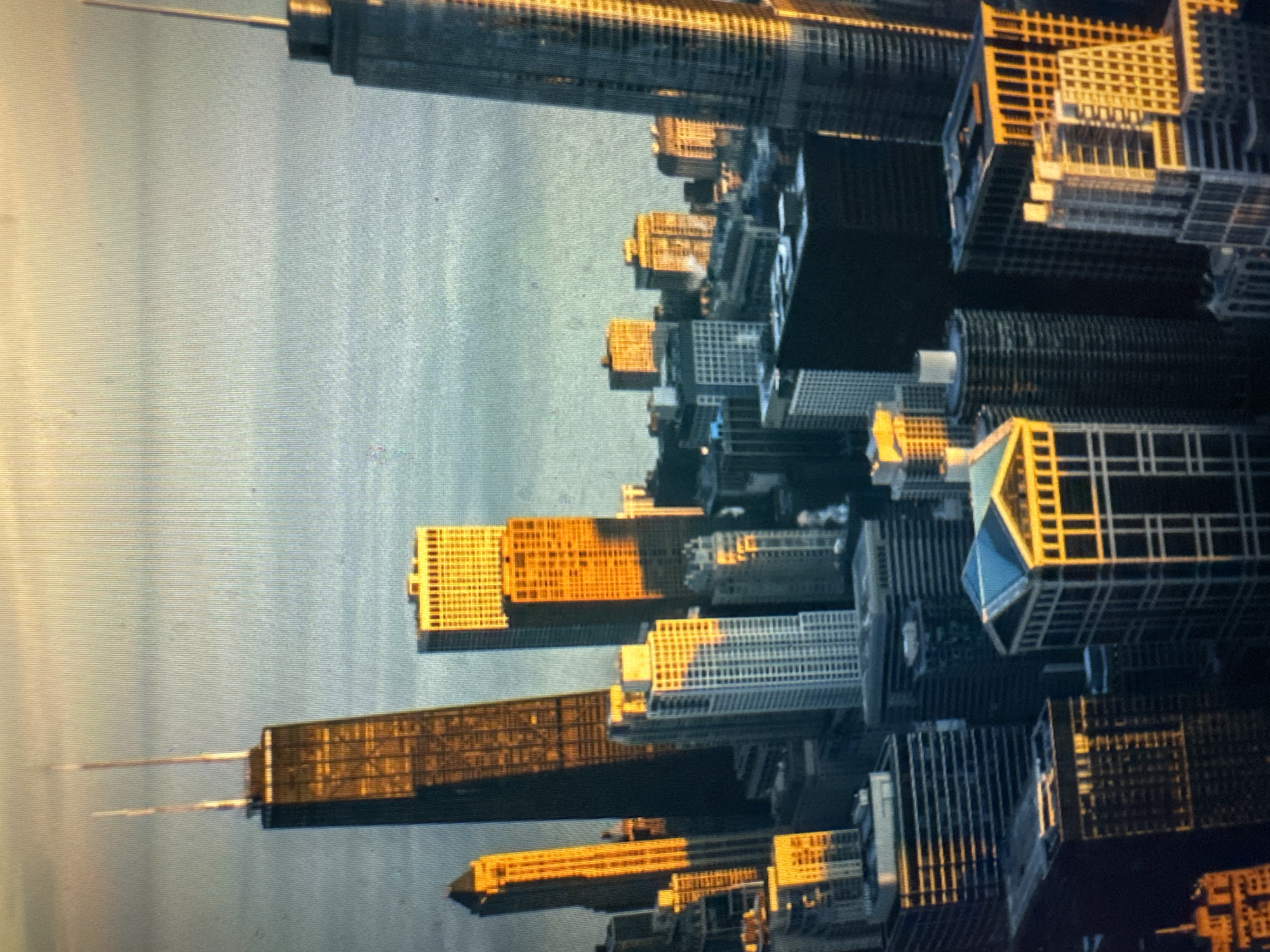The image shows a cityscape with several skyscrapers at an unknown location. The buildings are of various heights and architectural styles, with some featuring glass facades and others with more traditional brickwork. The sun is low in the sky, casting long shadows and illuminating the buildings with a warm, golden light, suggesting either sunrise or sunset. The water in the foreground is calm and reflects the light, creating a serene atmosphere. No people are visible in the image, and the focus is solely on the urban landscape. The overall impression is one of grandeur and the impressive scale of modern architecture.