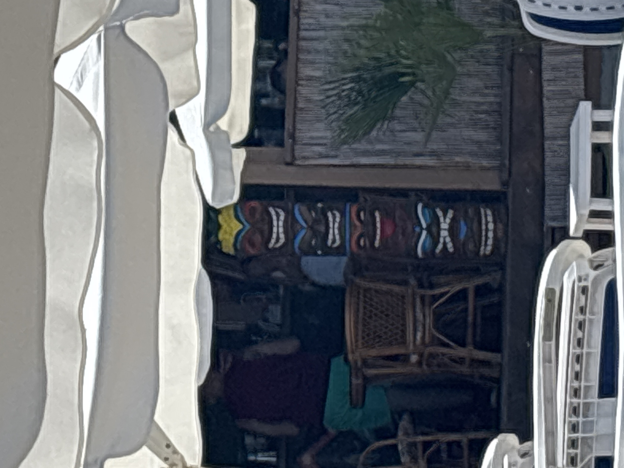 The image depicts an outdoor scene, likely a beach bar or resort area, with a tropical aesthetic. In the foreground, a white, modern lounge chair with blue accents is visible. Behind it, a low white table sits on a dark surface. Further back, a row of woven chairs and a small table are present. A person wearing a dark sleeveless top and green shorts is partially visible to the left, standing near a bar area.
The main focal point, centrally located, is a tall tiki totem pole featuring multiple carved faces with distinct expressions and colorful headdresses. The totem is positioned in front of a wall constructed of vertical bamboo or reed-like material. To the right of the totem, a tropical plant with green fronds adds to the ambiance. The scene is partially covered by a white canopy or umbrella, creating shade. The lighting suggests it is daytime. The background appears to be a building or structure associated with the outdoor establishment. The overall atmosphere is relaxed and summery.